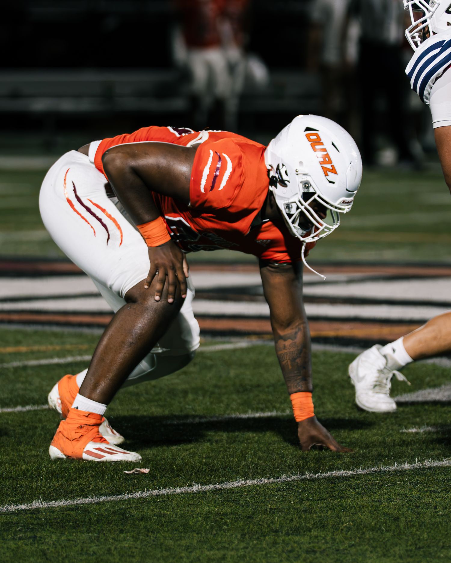 Football player in orange uniform crouches in defensive stance on field at night game.