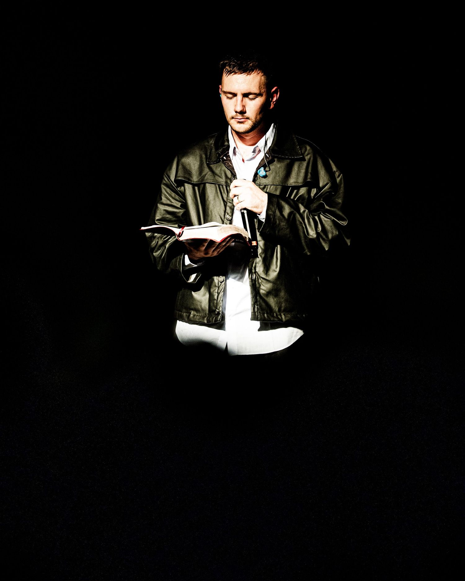 A man in a dark leather jacket reads a book under dramatic spotlight lighting against a black background.