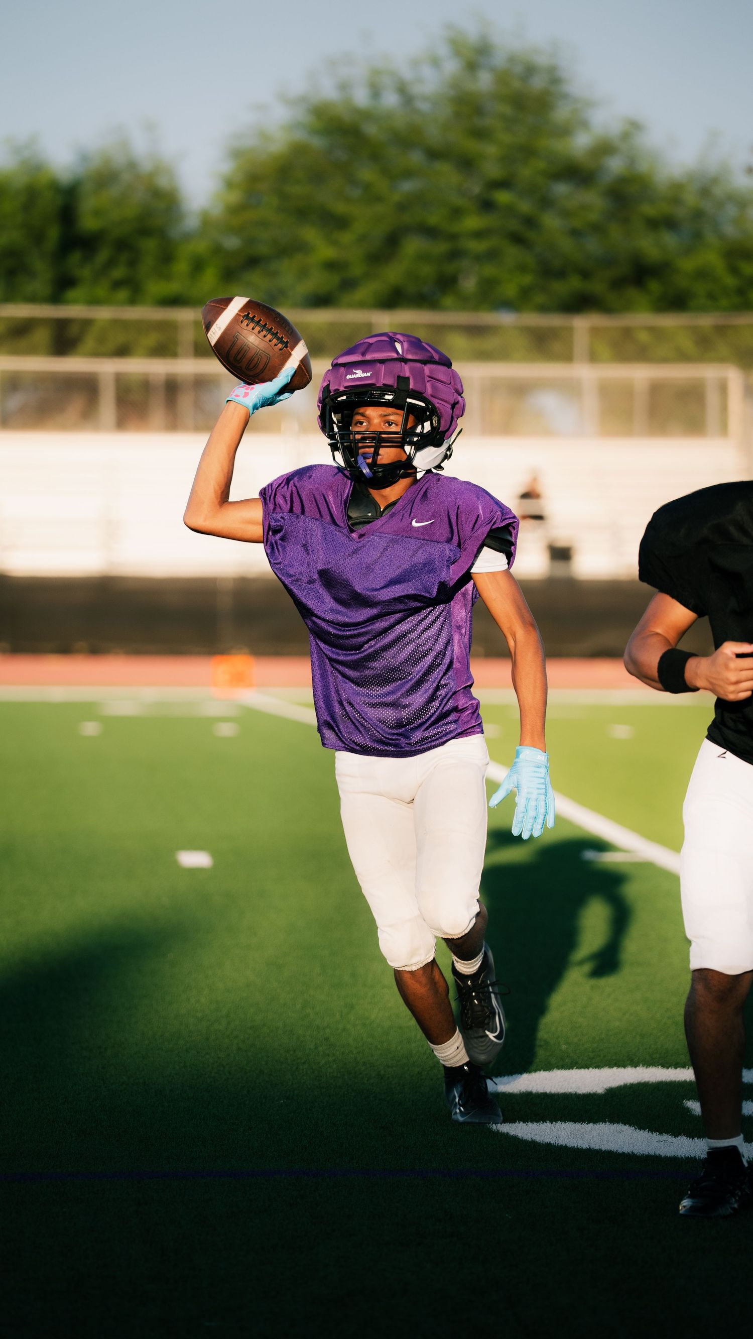 Youth football player in purple Nike jersey throws a football during practice on a green turf field.
