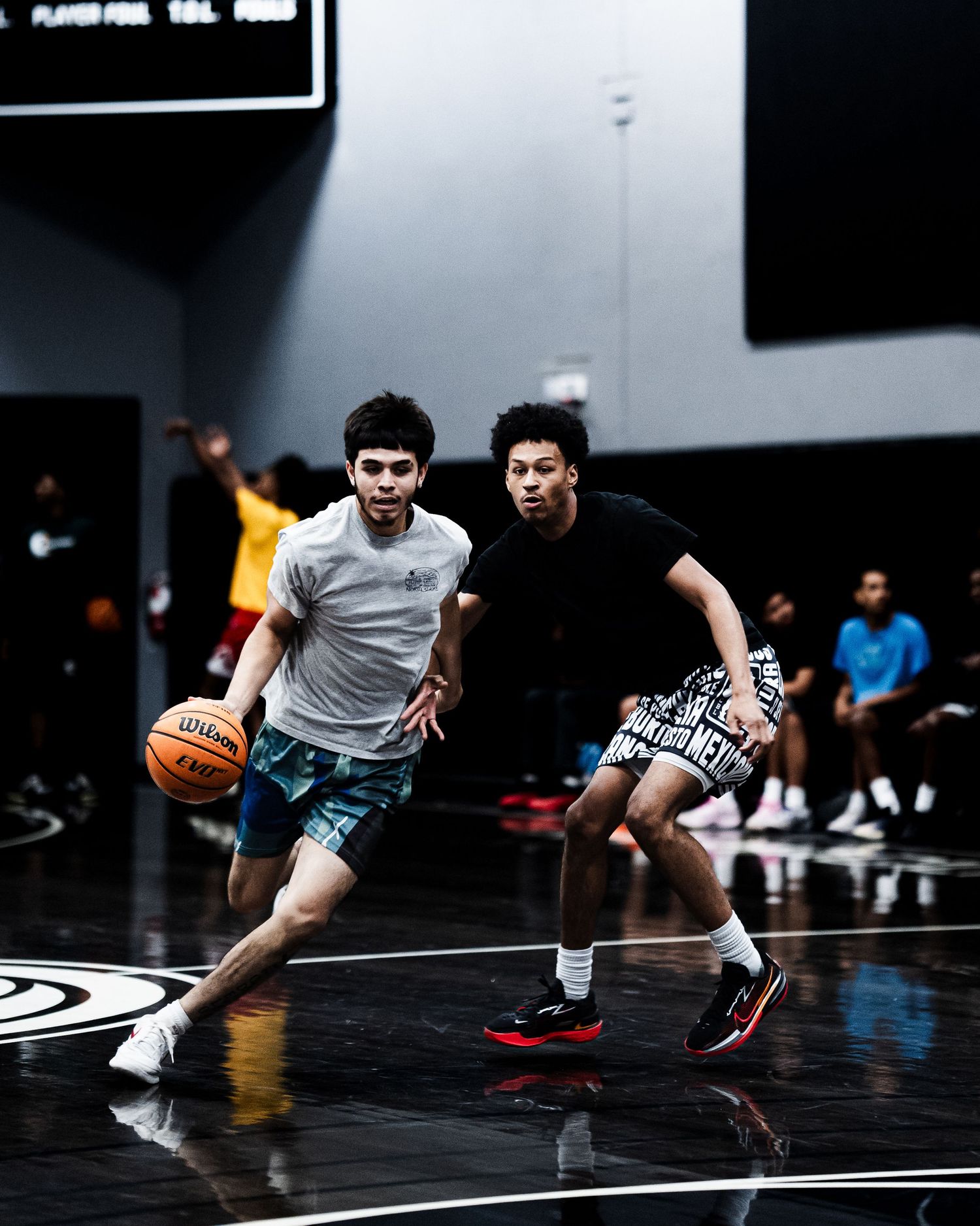Two young basketball players competing in a dribble drive drill on a shiny indoor court.