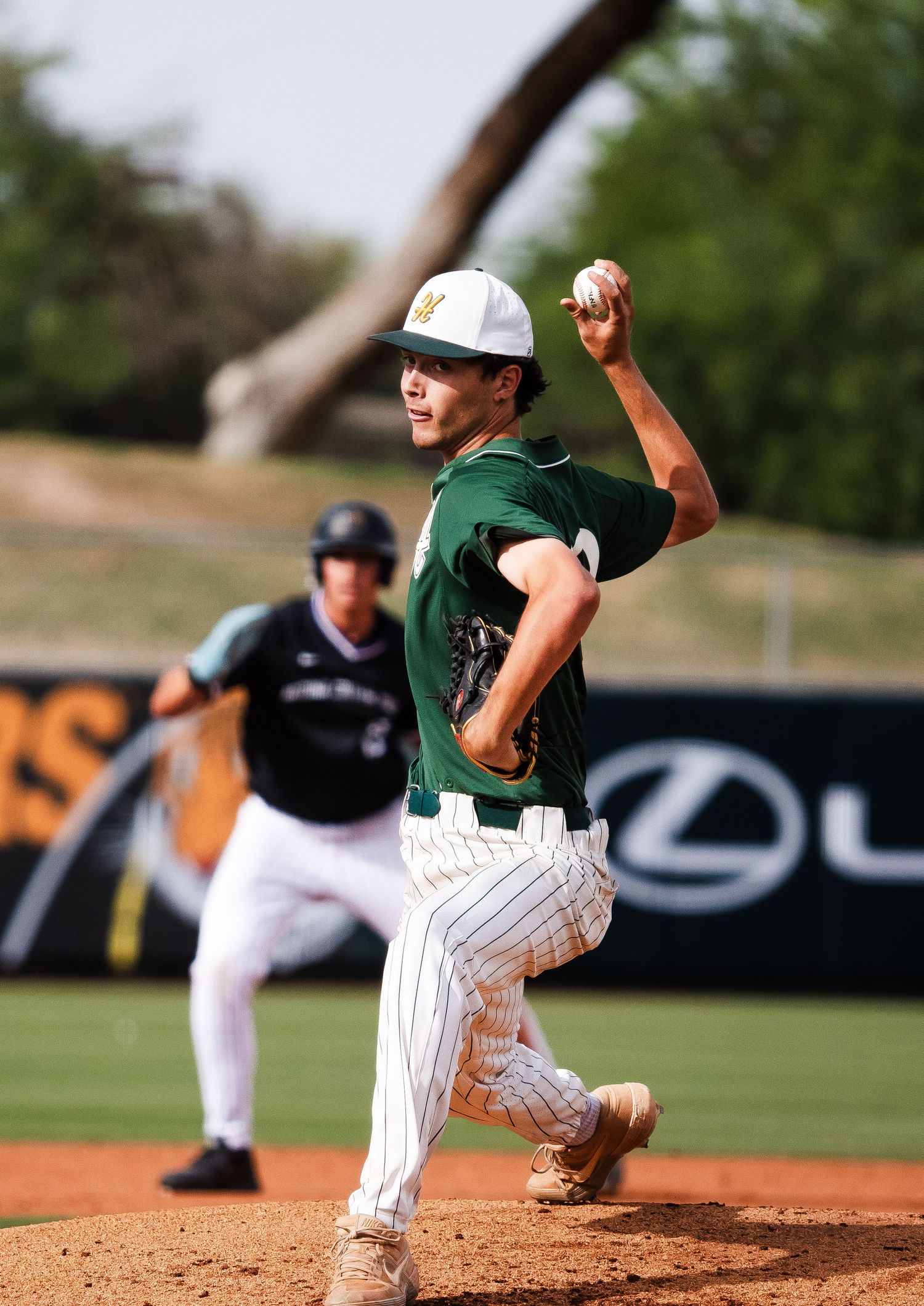 Baseball pitcher in green uniform mid-throw on mound, with baserunner and Lexus advertisement visible in background.