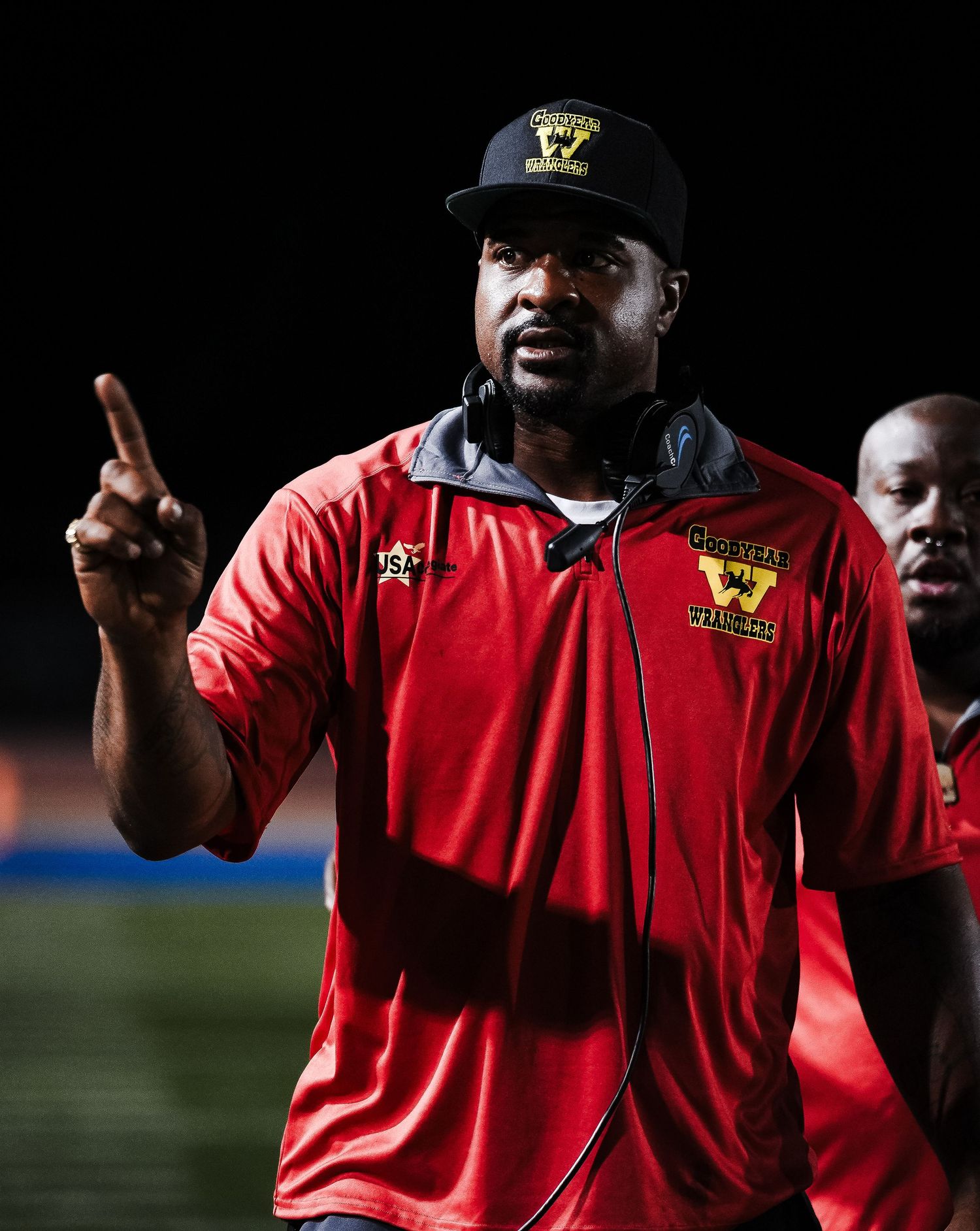 A football coach in a red jersey and black cap gestures on the sideline during a night game.