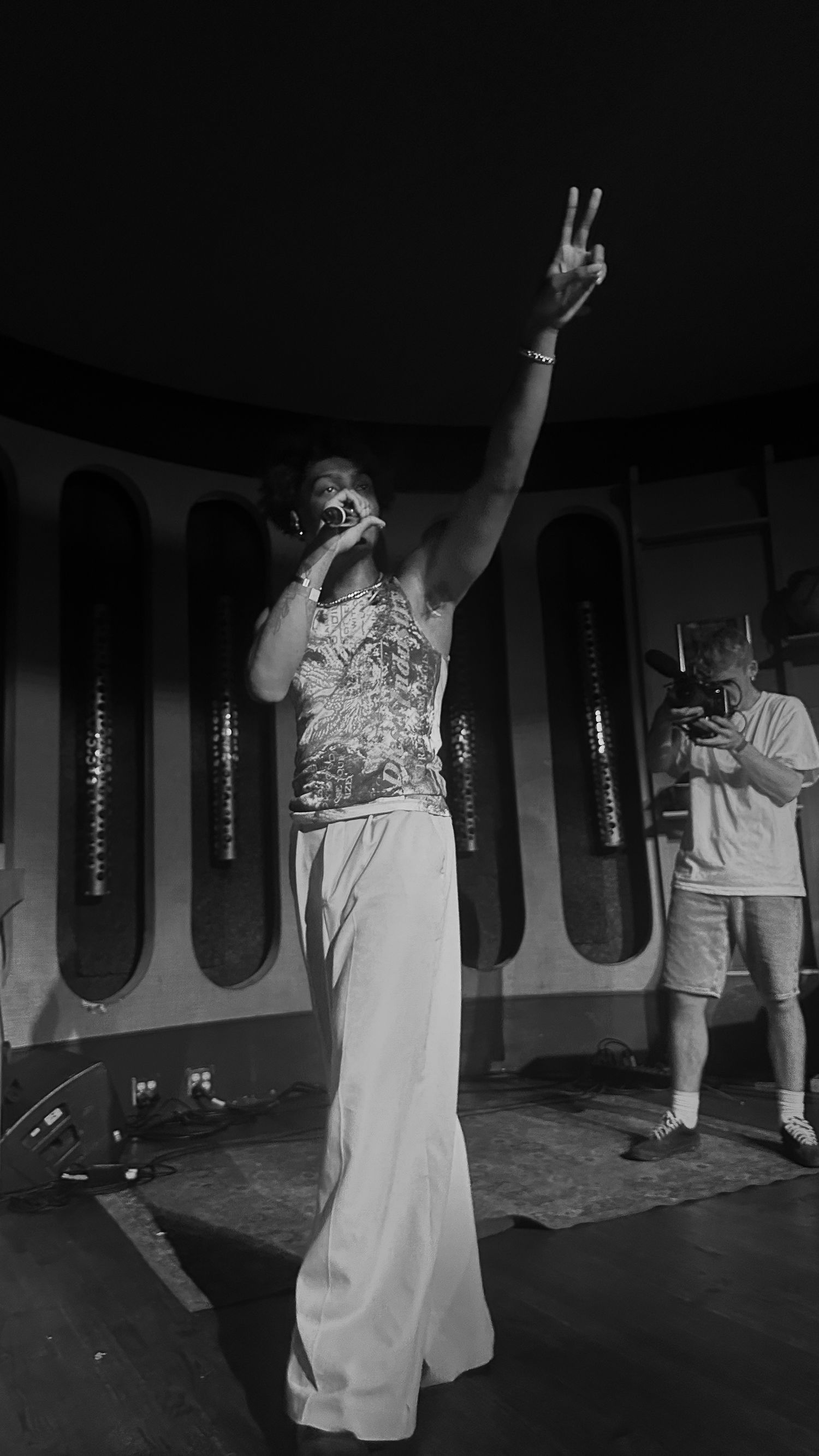 Black and white photo of a performer on stage holding a mic, raising a peace sign, with a photographer in the background.