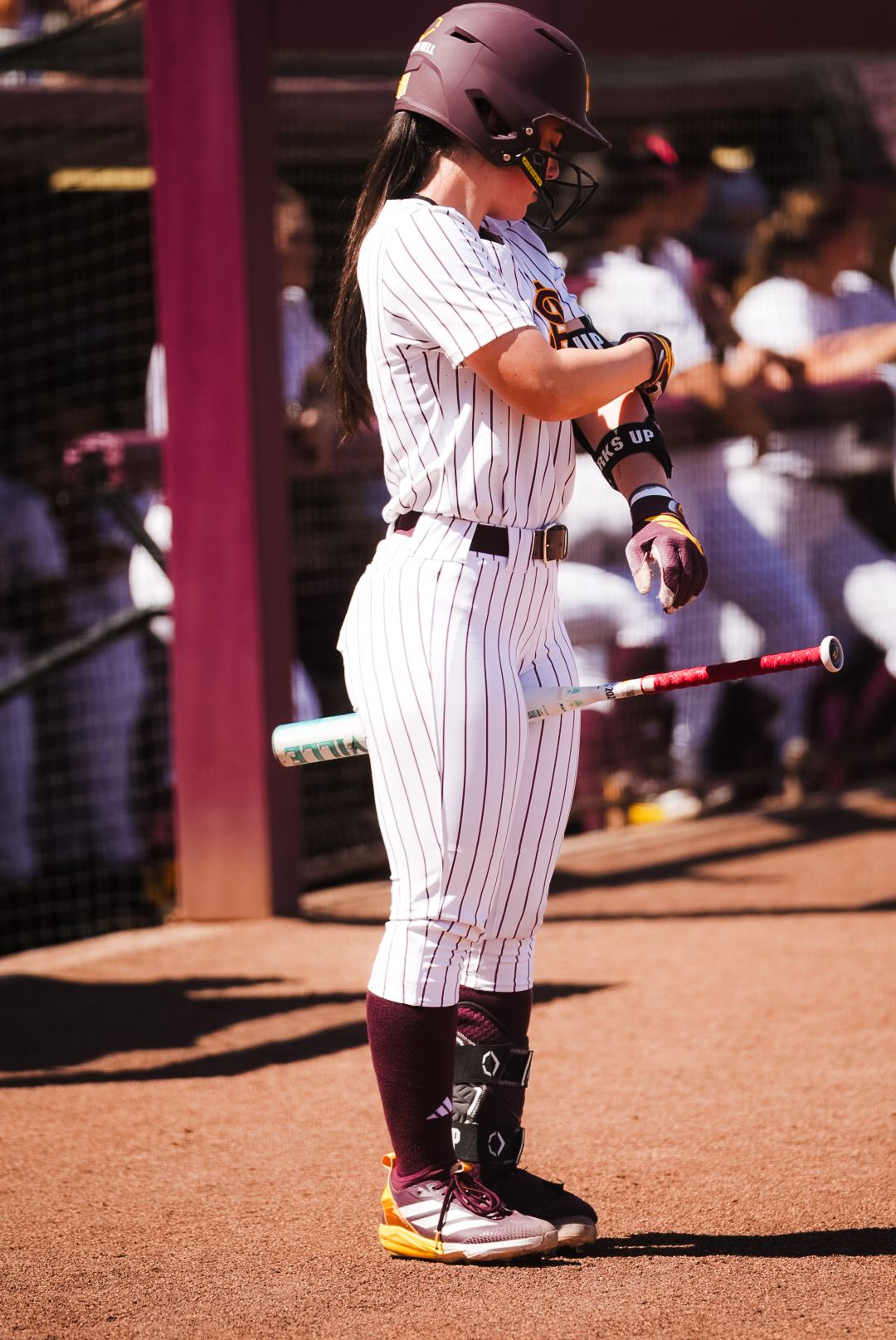 Female softball player in maroon pinstripe uniform holds bat while preparing to bat during a college softball game.