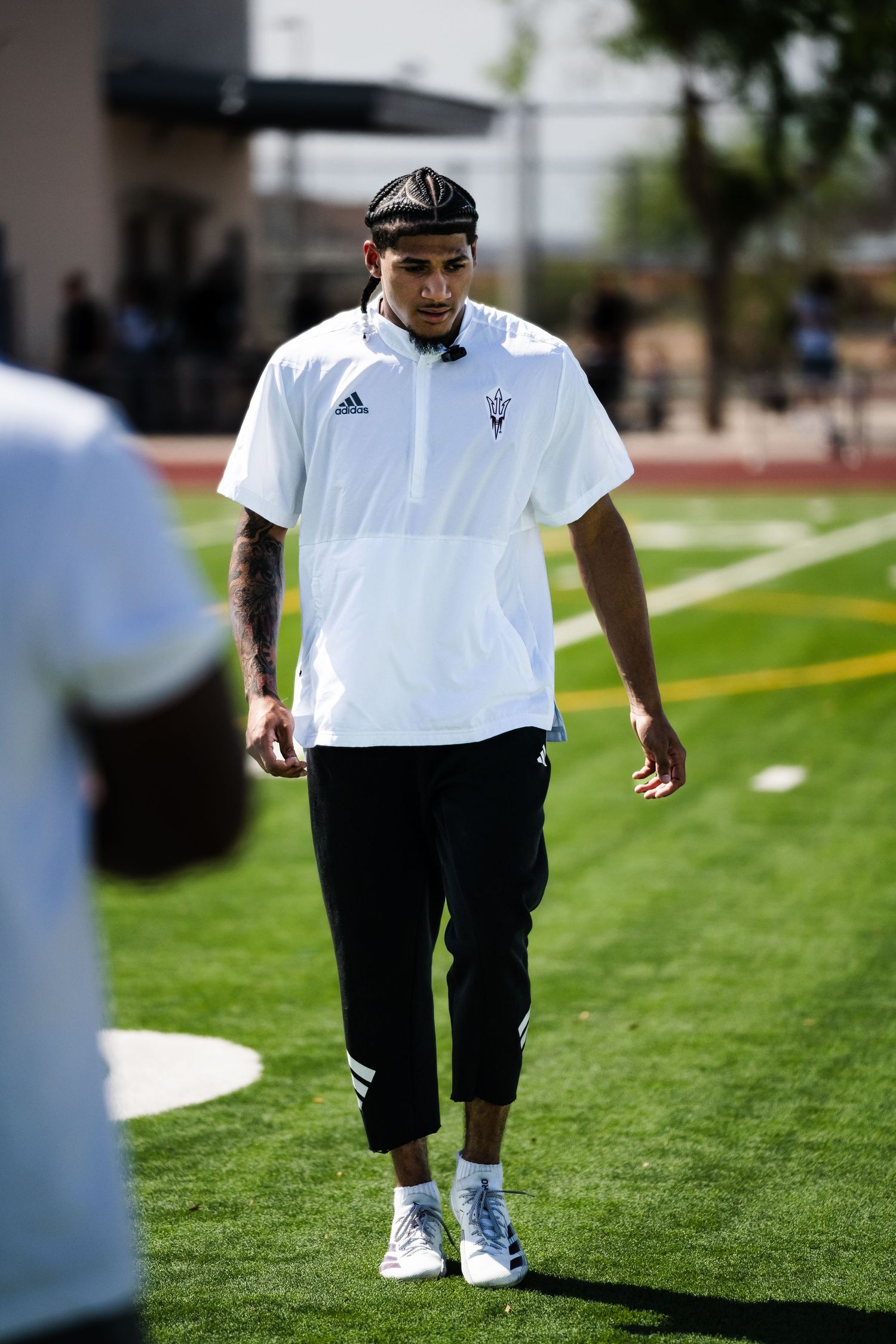 Tattooed man in Adidas Arizona State white shirt walks on green turf football practice field.