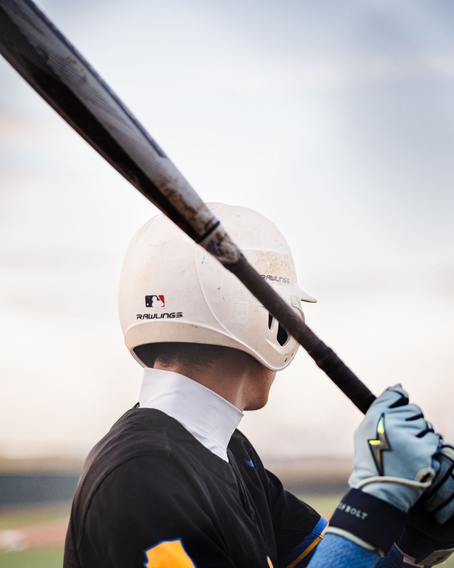 Baseball batter wearing a white Rawlings MLB helmet holds a black bat over shoulder, ready to swing.