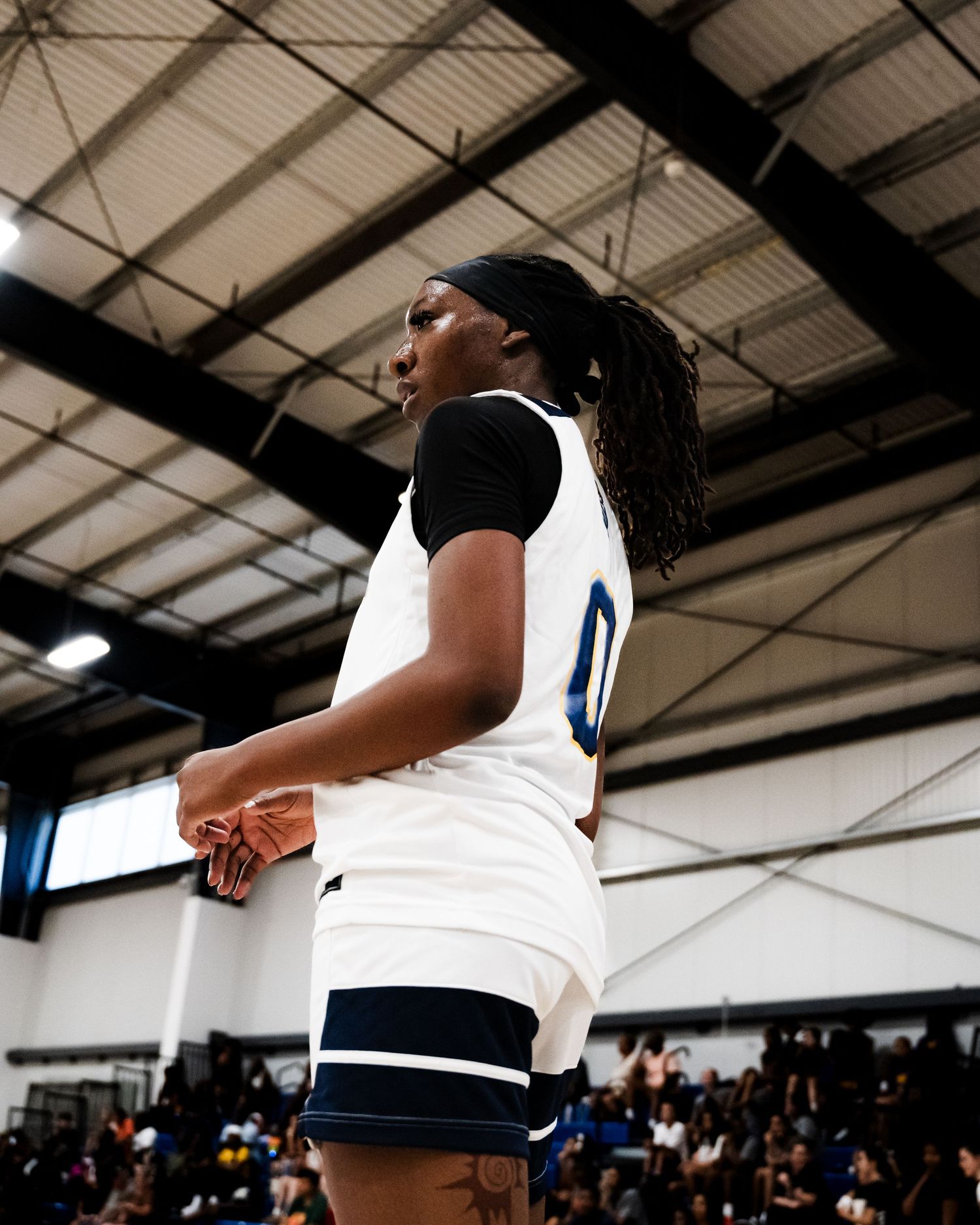 A female basketball player wearing a white number 0 jersey stands on the sideline of an indoor gym.