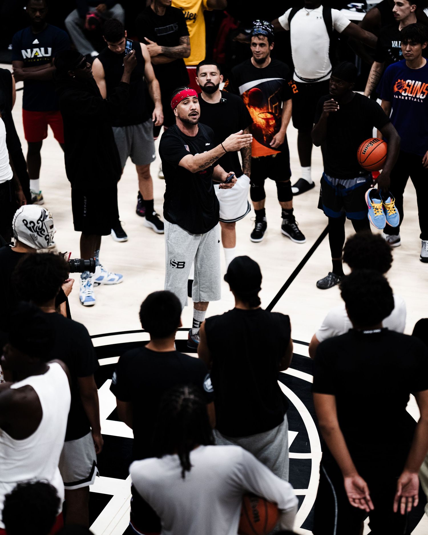 Coach in black shirt with red headband speaks to a large group of basketball players on an indoor court.