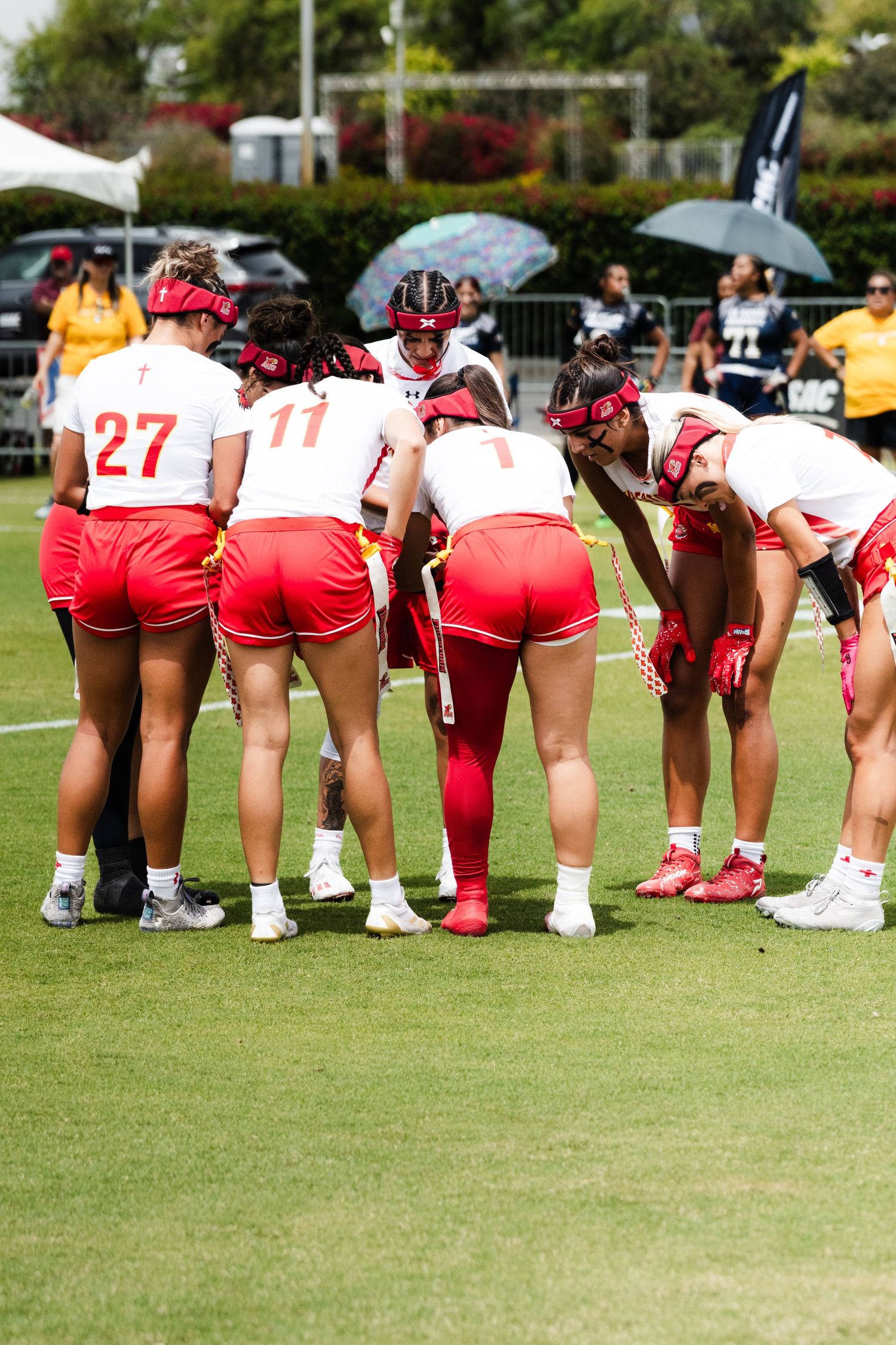 Women's flag football team in red uniforms huddling together on green field during sunny outdoor game.