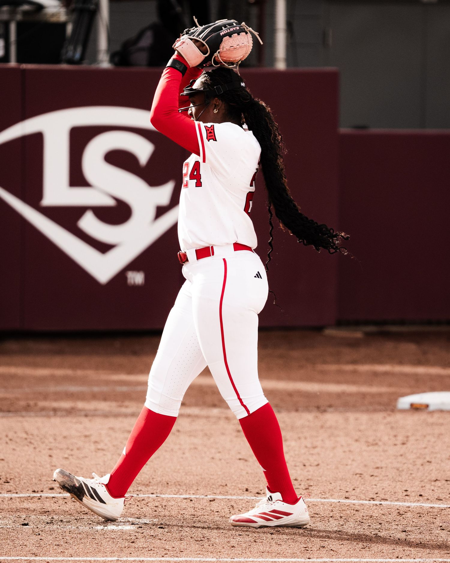 Softball pitcher in white and red uniform winds up to throw on a dirt mound with Louisville Slugger signage behind.