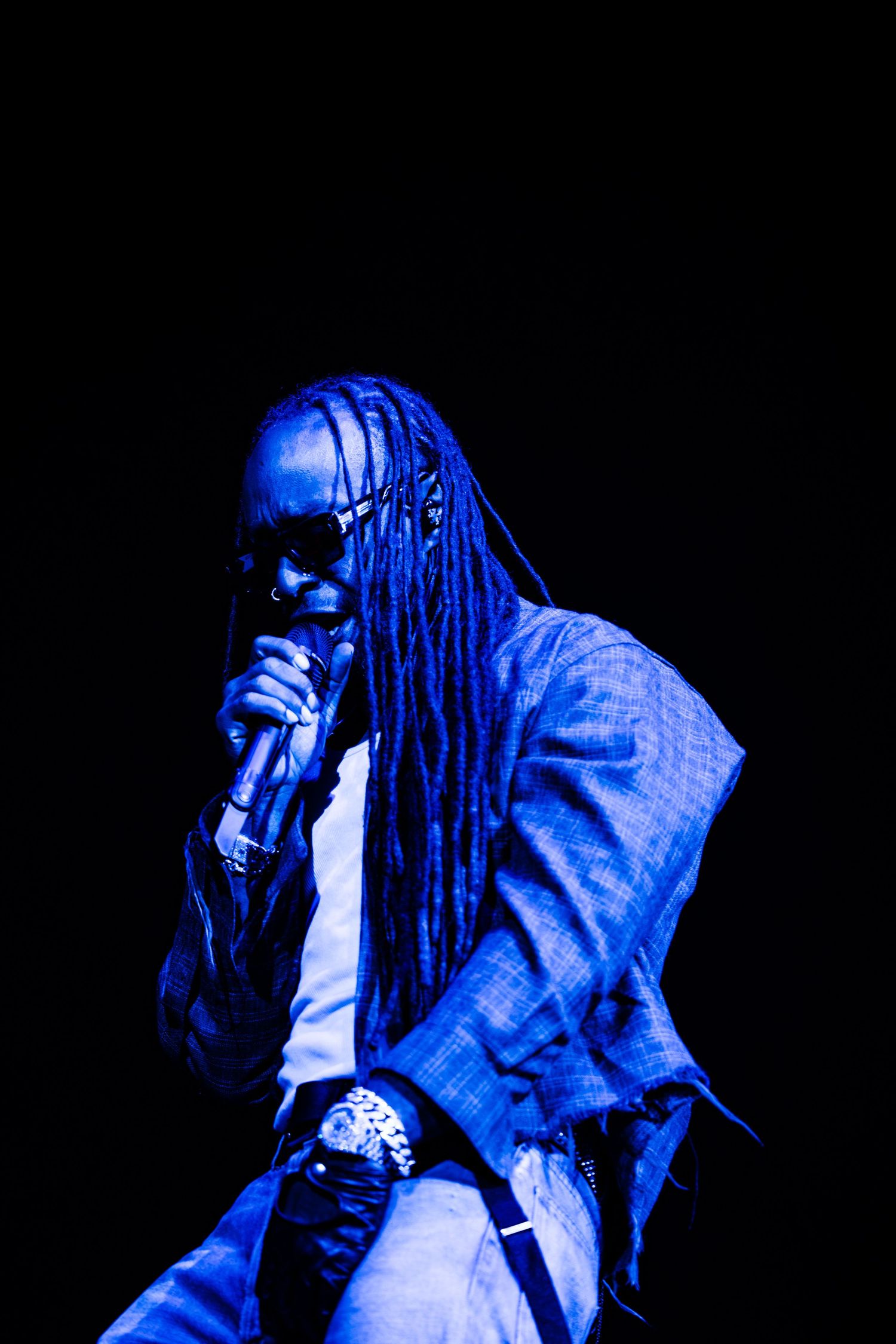 Artist with long dreadlocks and sunglasses sings into a microphone, wearing a plaid jacket and watch, bathed in deep blue stage lighting.