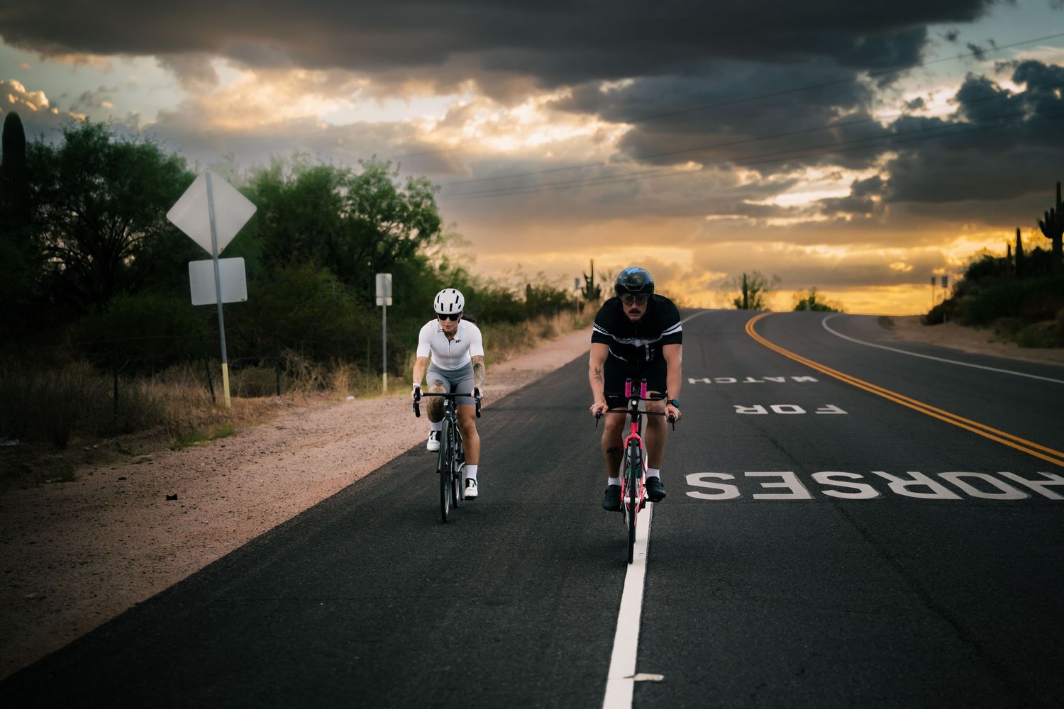 Two cyclists ride road bikes on a scenic desert highway at sunset with dramatic storm clouds overhead.