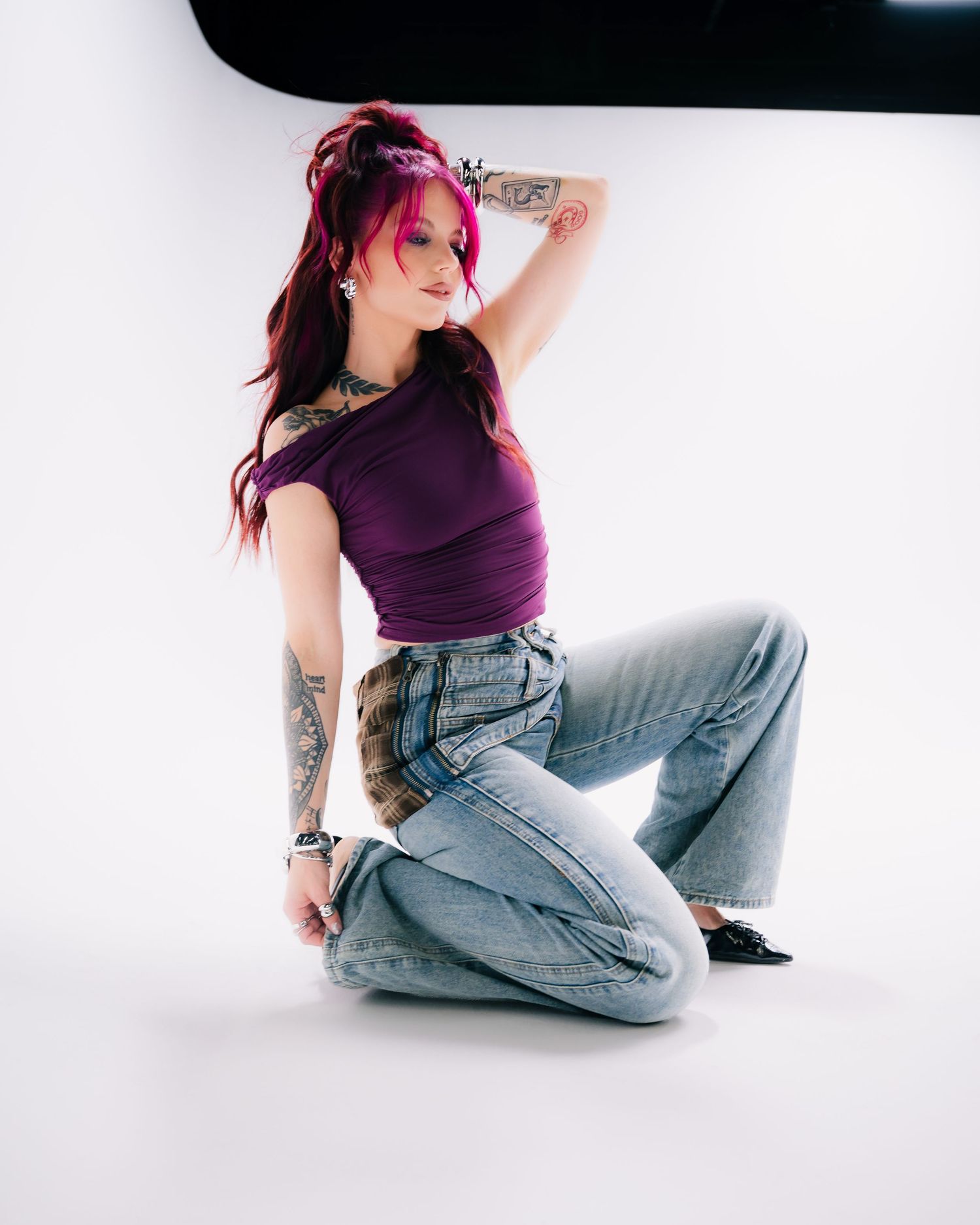 Woman with pink hair and tattoos poses in purple crop top and wide-leg jeans on white studio background.
