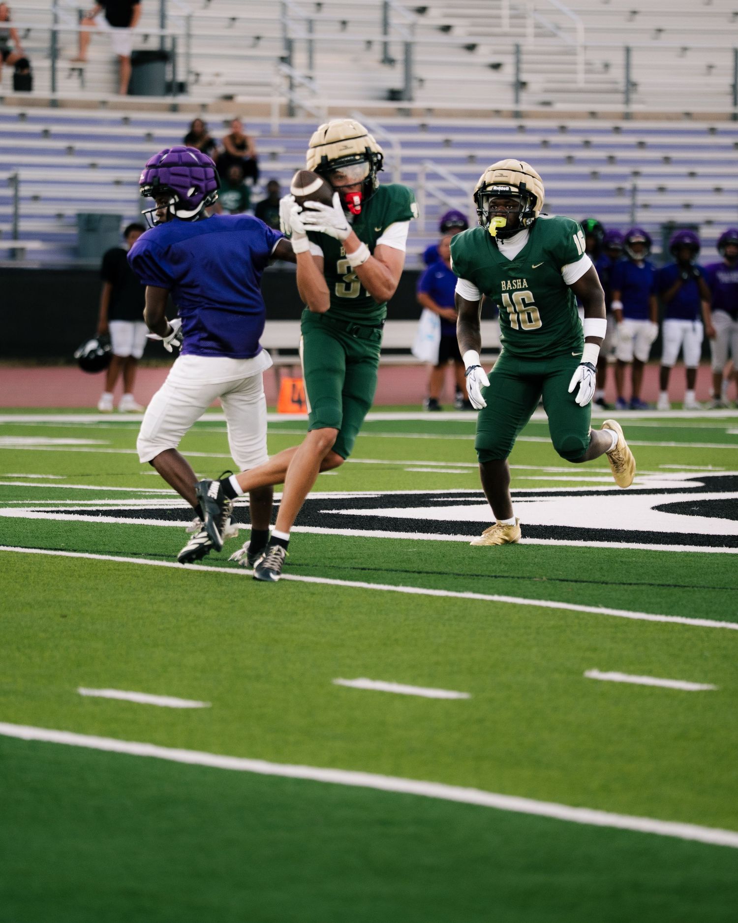 High school football players in green and purple uniforms compete on a turf field during a game.