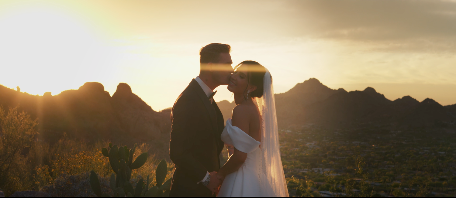 Couple sharing a romantic kiss at sunset with stunning mountain silhouettes in the background at an outdoor wedding.