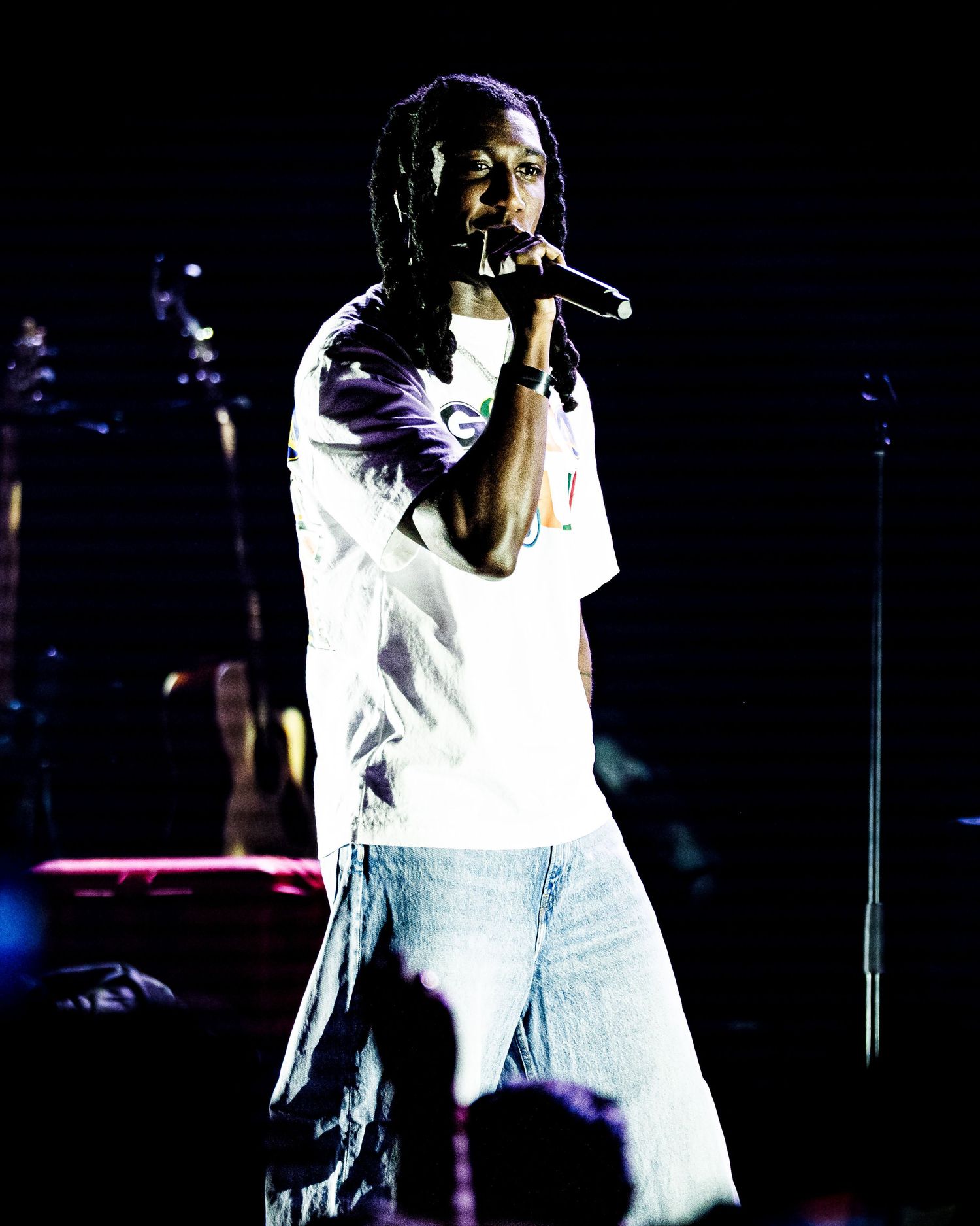 Dreadlocked hip-hop artist in white t-shirt performs with microphone under purple and blue stage lights.