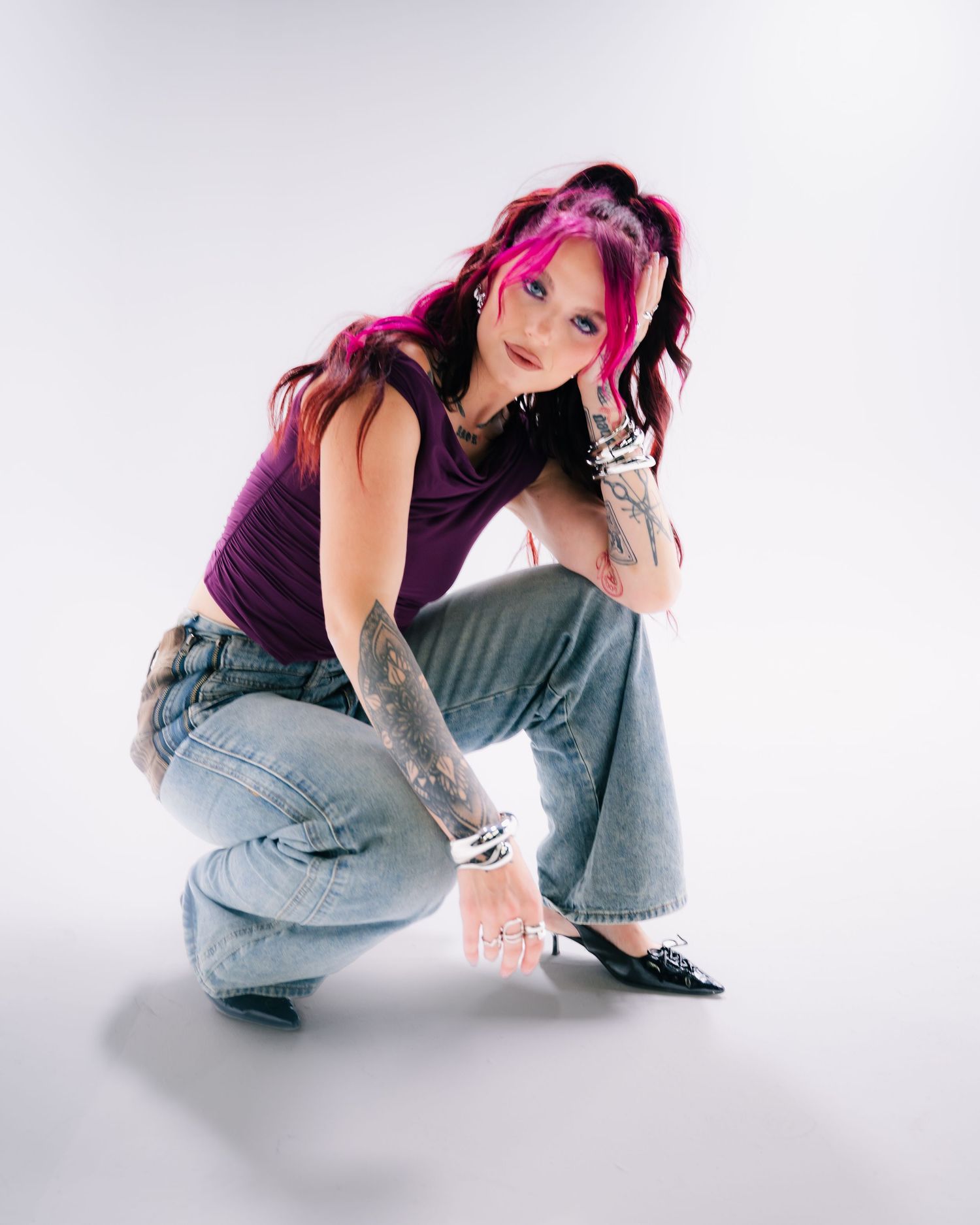 Woman with pink hair and tattoos poses crouching in jeans and purple top against white background.