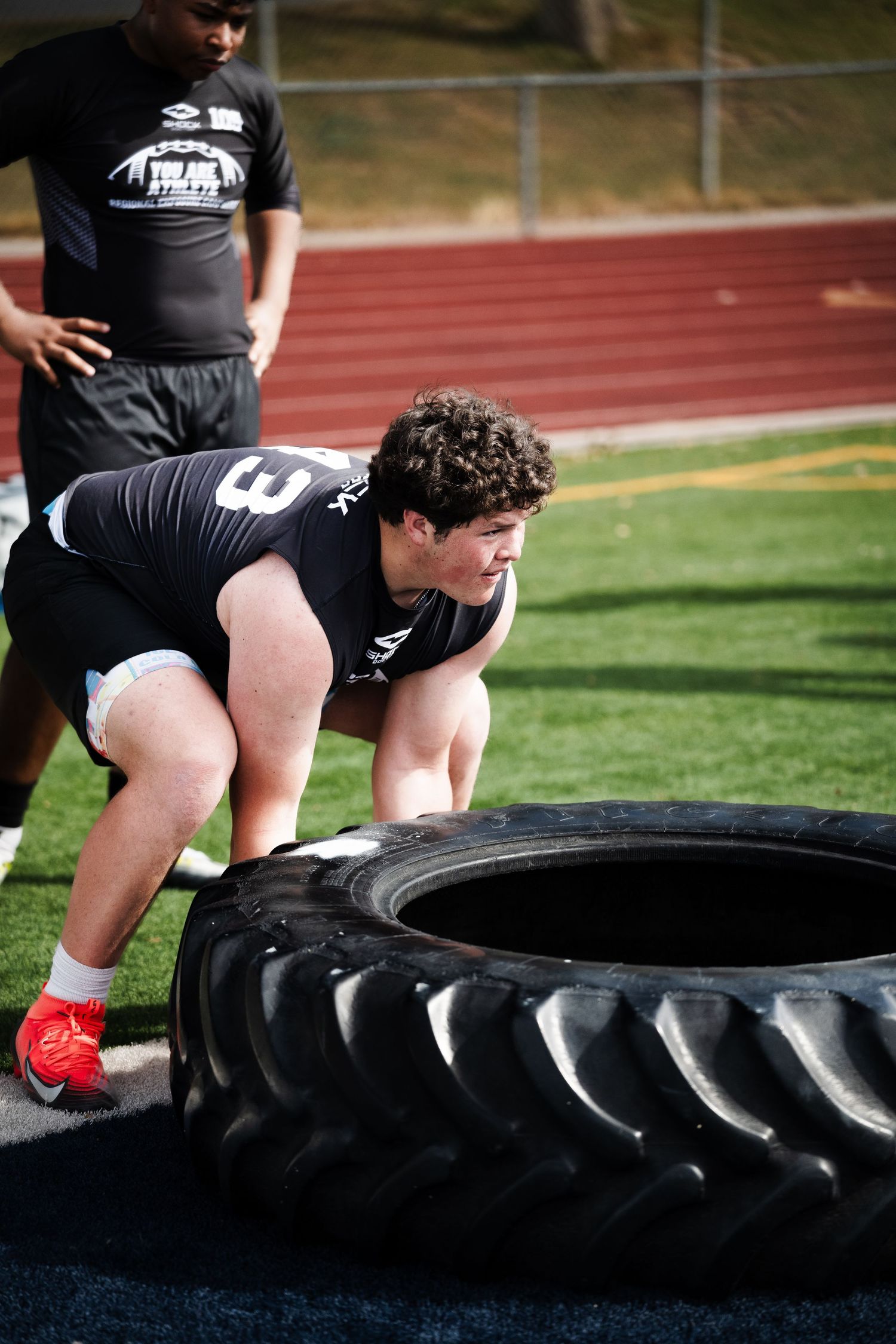 Young football player in black jersey pushing heavy tractor tire during combine strength drill.