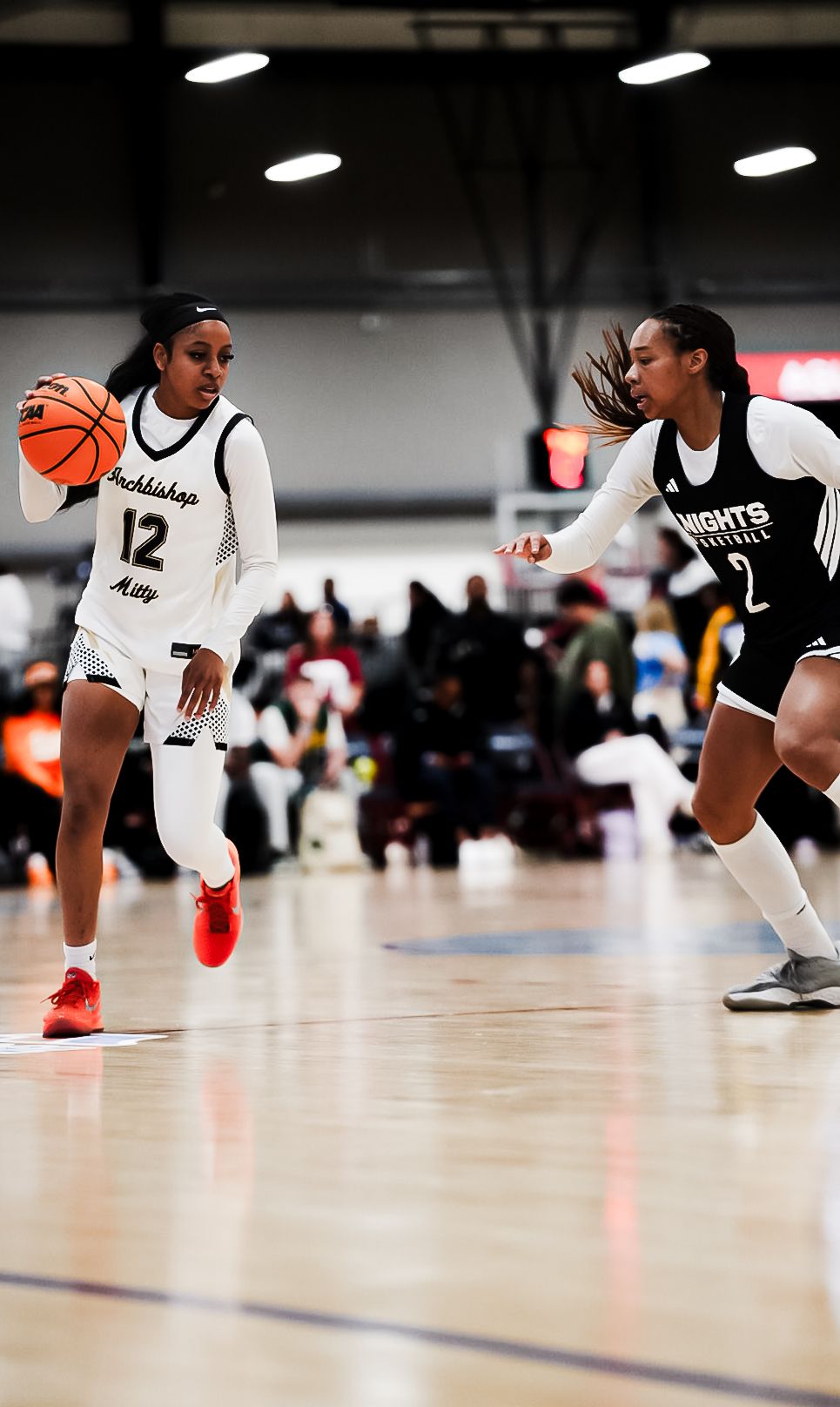 Player #12 in white Archbishop jersey dribbles basketball past defender in black Knights uniform on indoor court.
