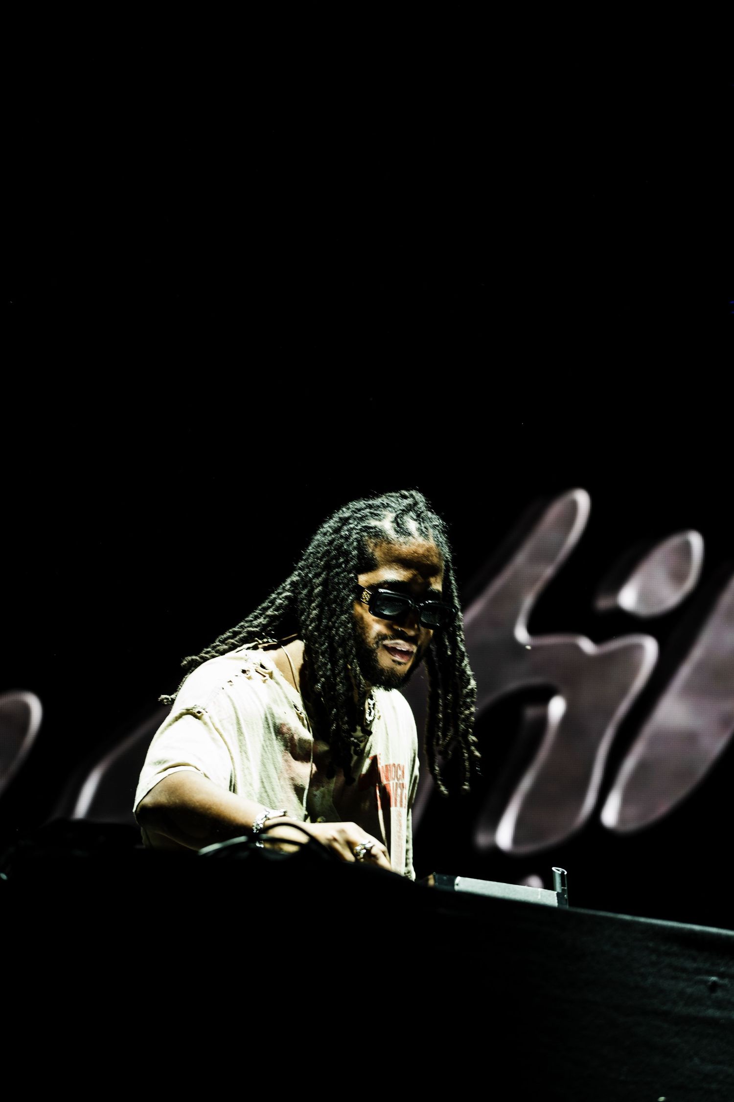 DJ with long locs and dark sunglasses performs at a mixing console on a dark stage with large glowing lettering behind.