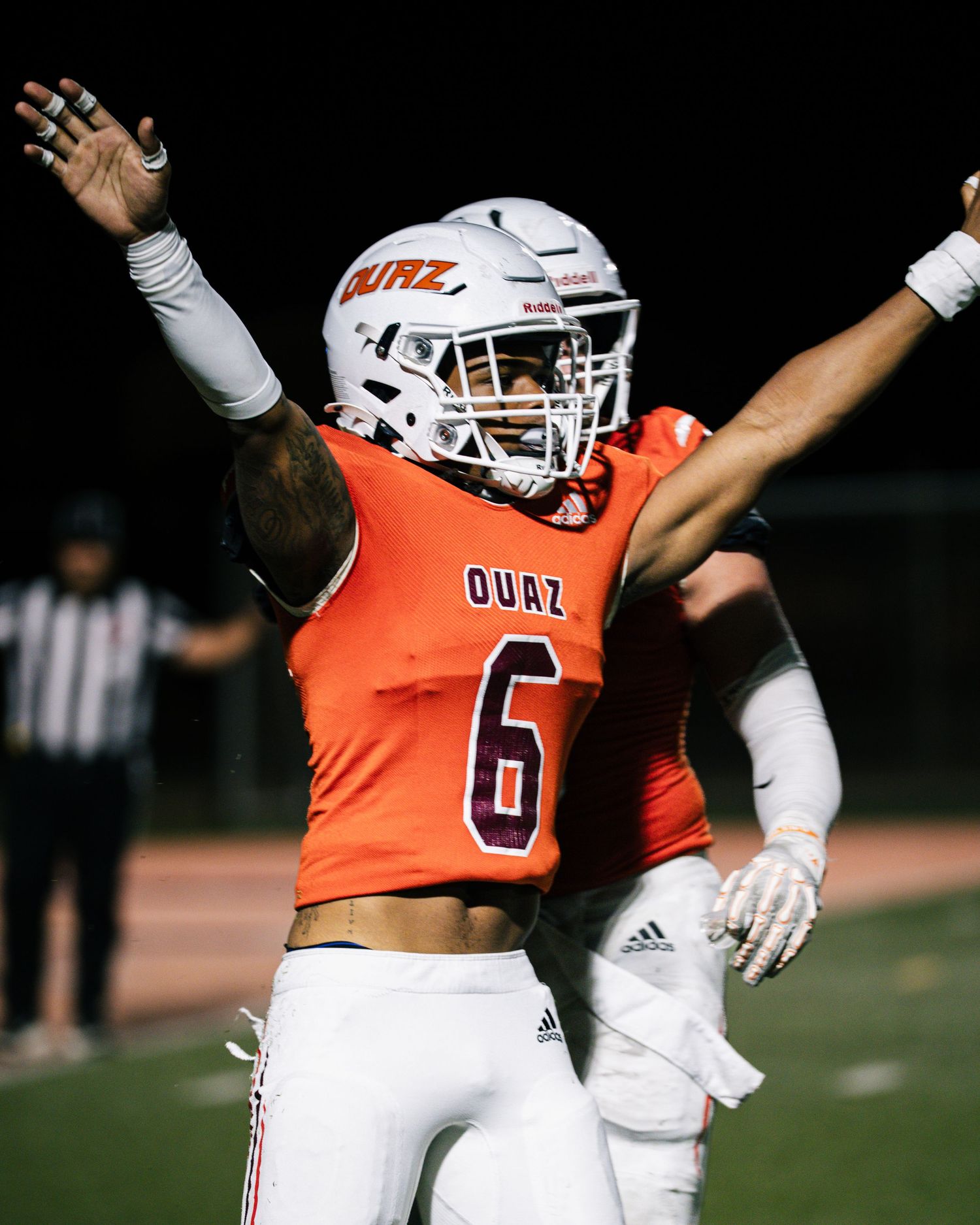 OUAZ football player #6 in orange uniform celebrates with arms raised during night game.