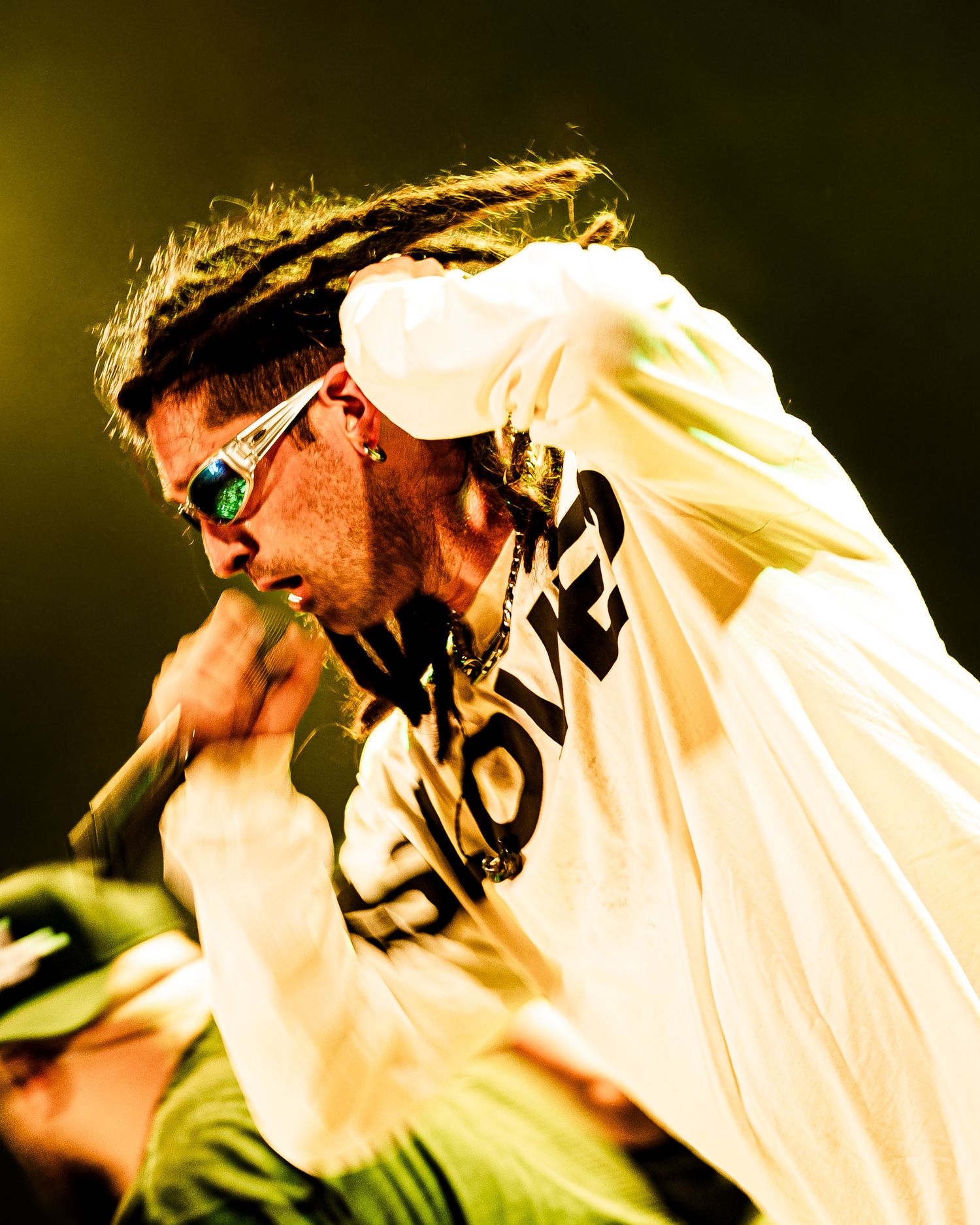 Energetic performer with dreadlocks and sunglasses singing into a microphone on stage in a white graphic tee.