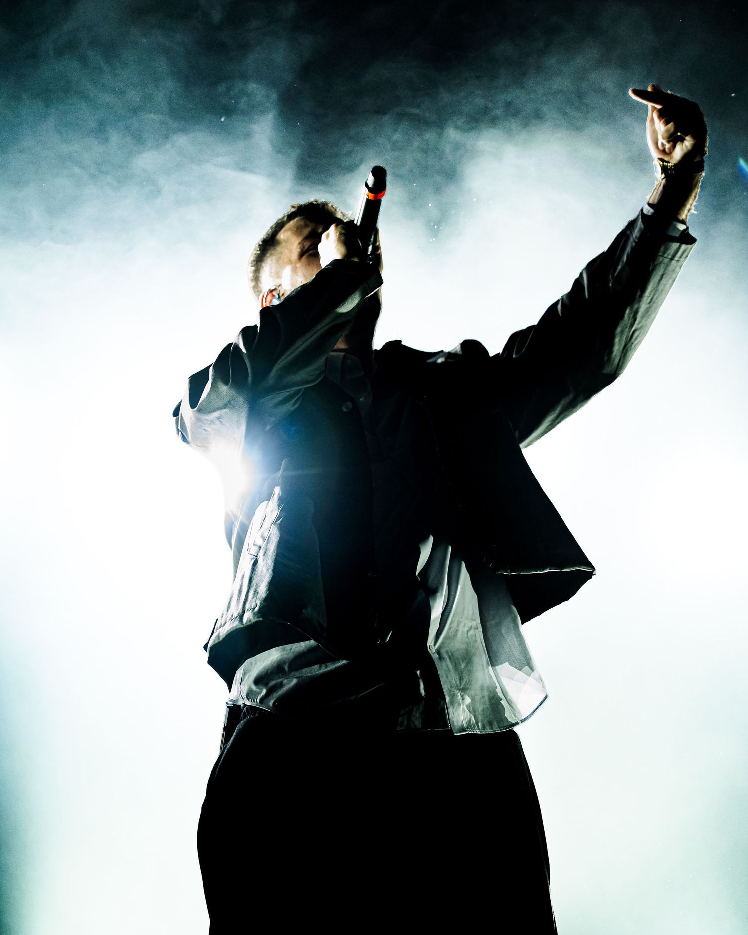 Dynamic concert performer holds microphone aloft in smoky backlit stage atmosphere during energetic live show.
