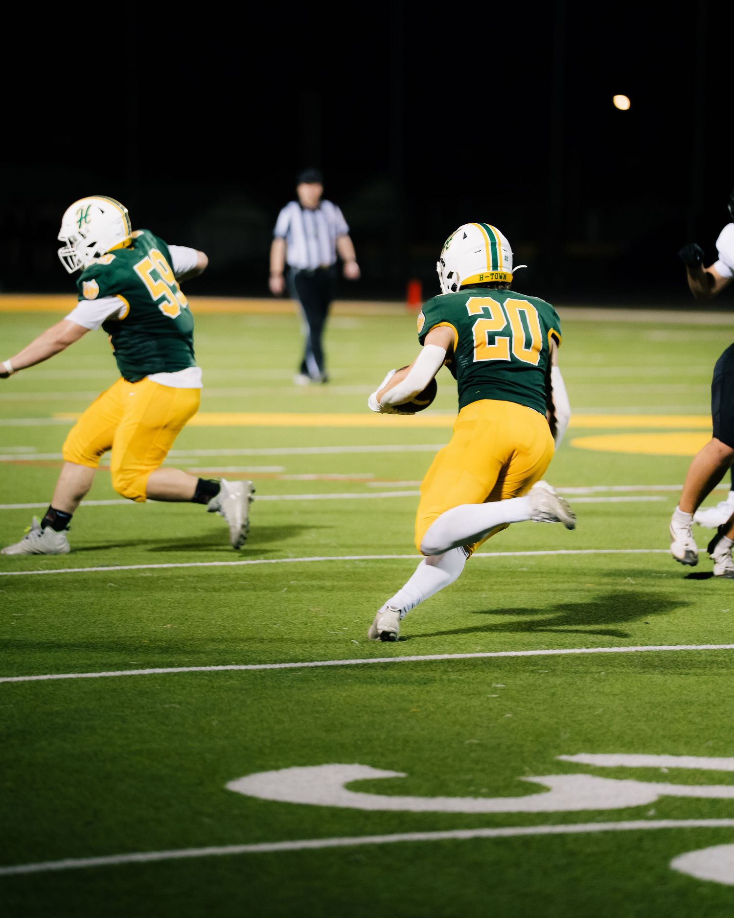 High school football players in green and gold uniforms run a play on a lit field during a night game.