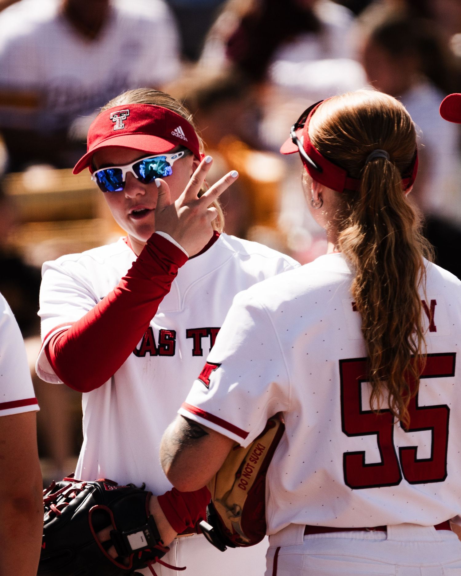 Texas Tech softball players in red and white uniforms discussing strategy during a game, player number 55 visible.