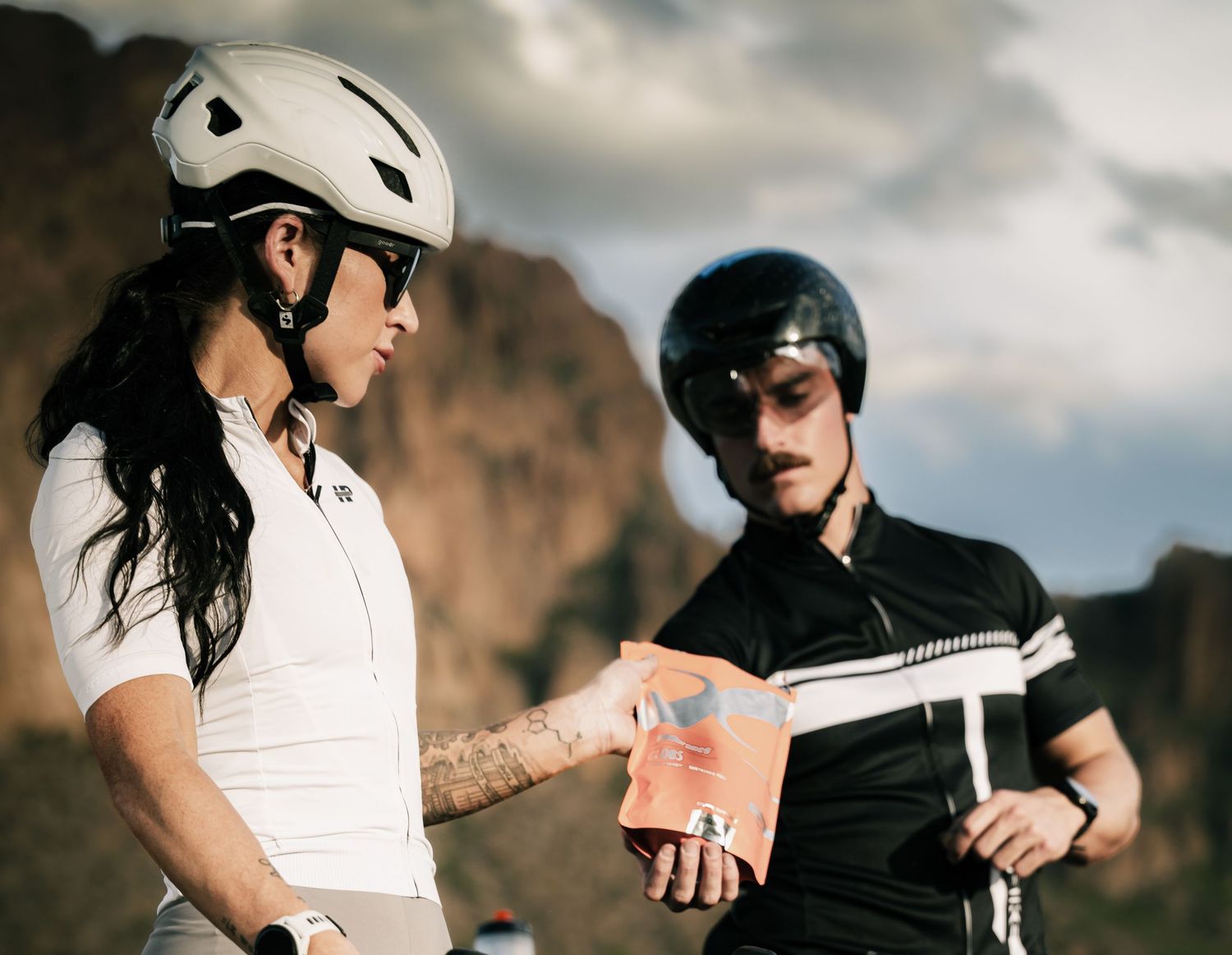 Two cyclists in helmets review a map together against a rocky mountain backdrop.