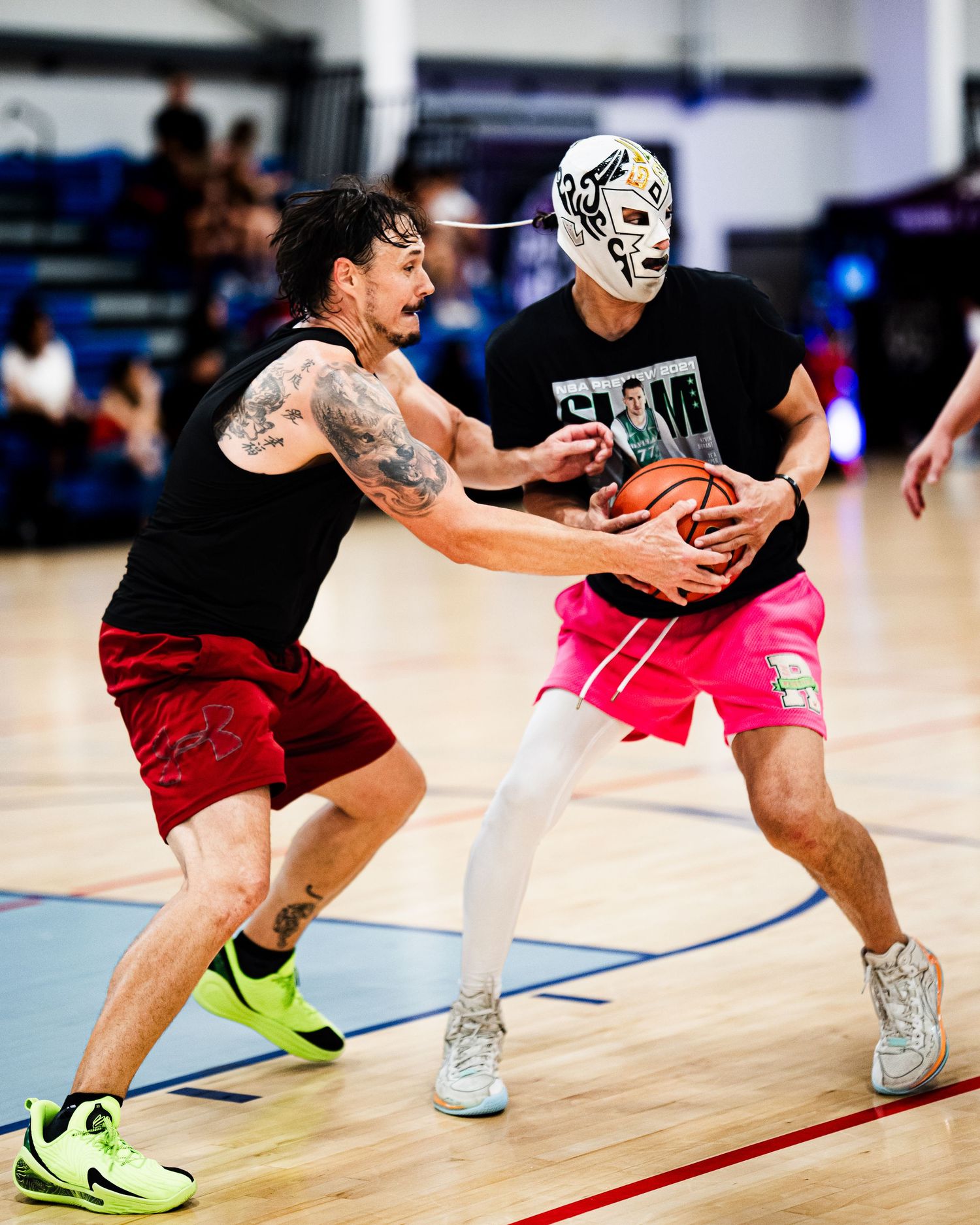 Two basketball players compete on indoor court, one wearing a lucha libre mask and pink shorts, the other with tattoos.
