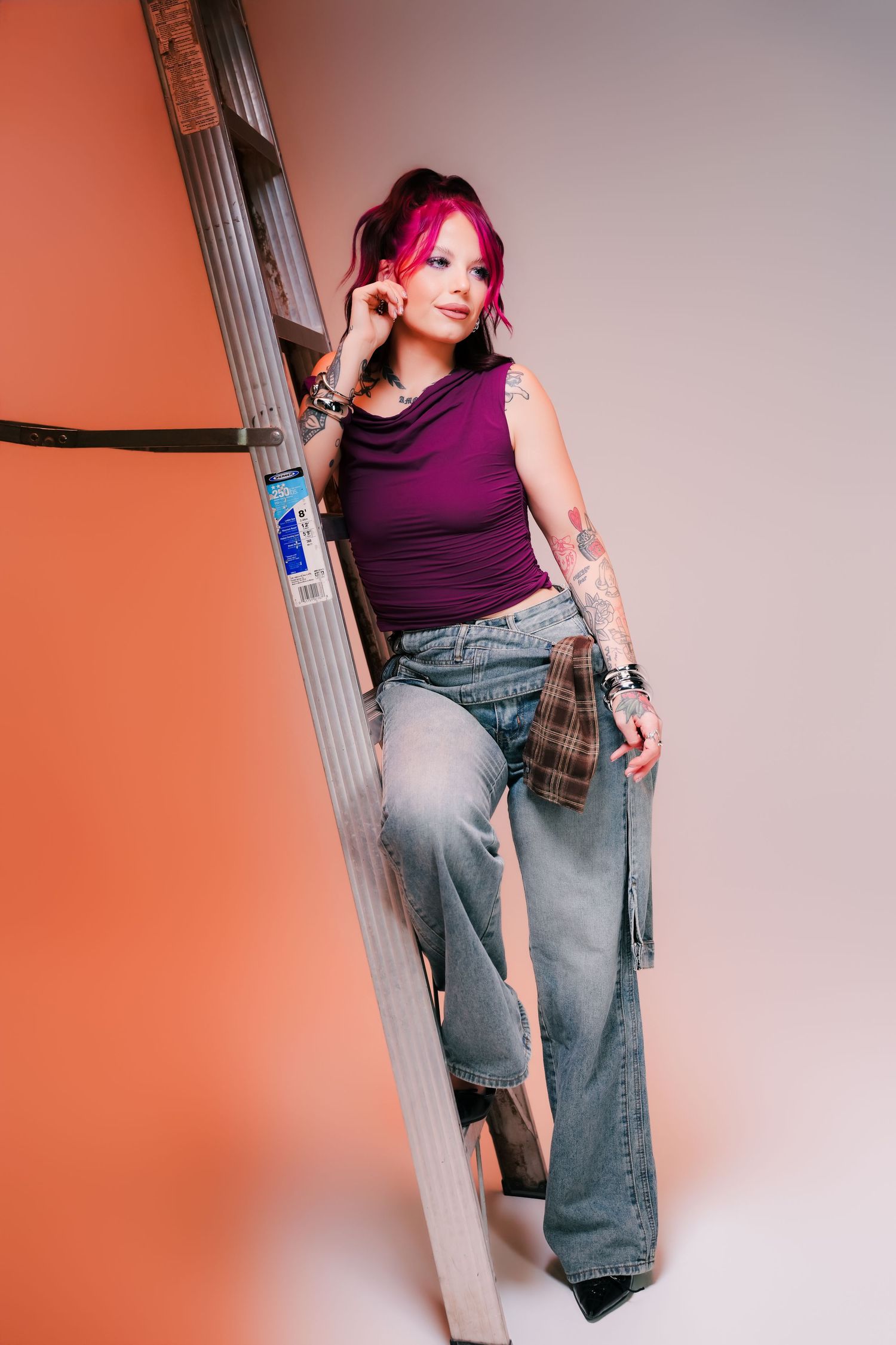 Woman with pink hair in purple top and jeans leans against a metal ladder, showcasing tattoos and layered bracelets.