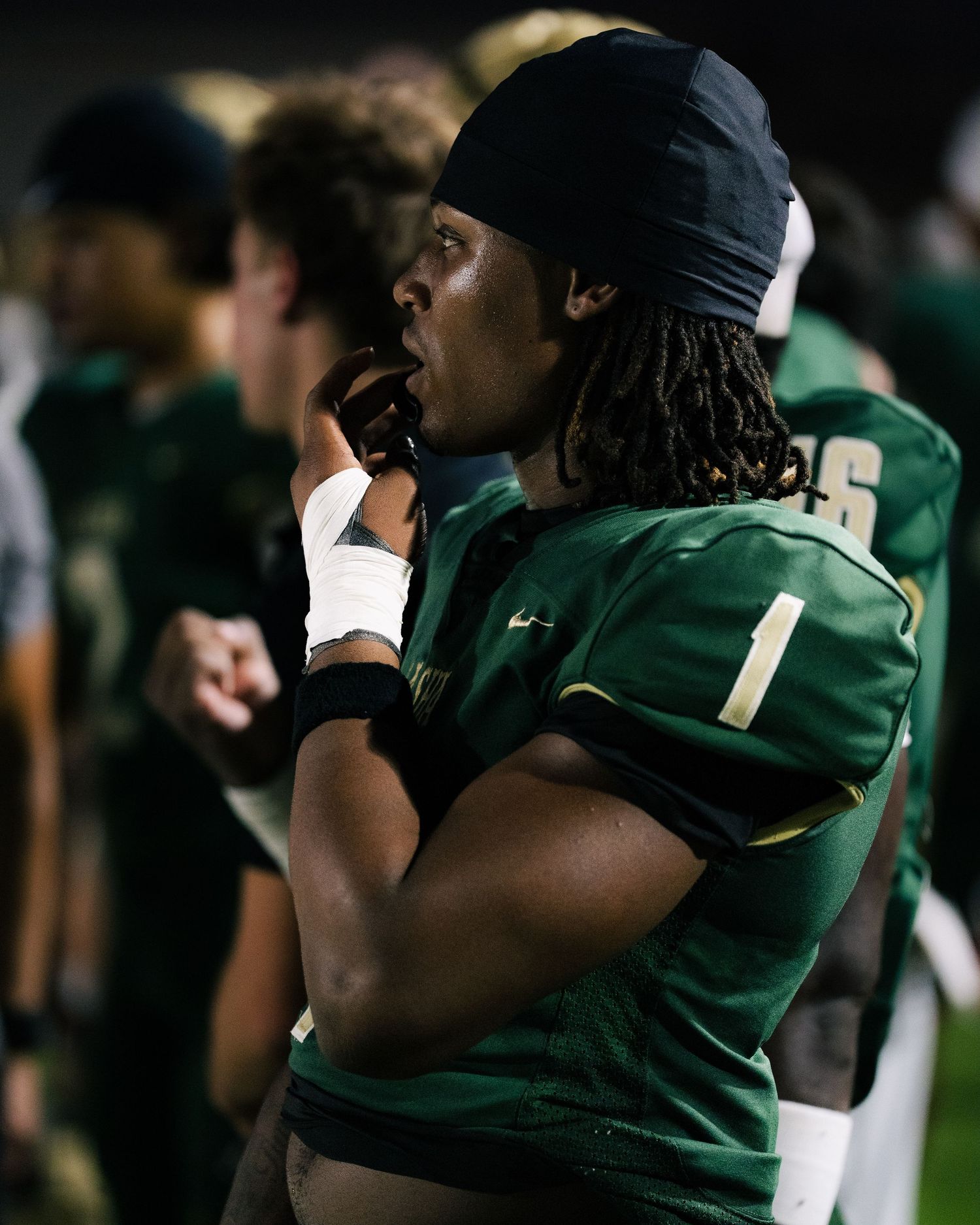 Football player wearing green jersey number 1 with dreadlocks and headband stands on sideline during night game.