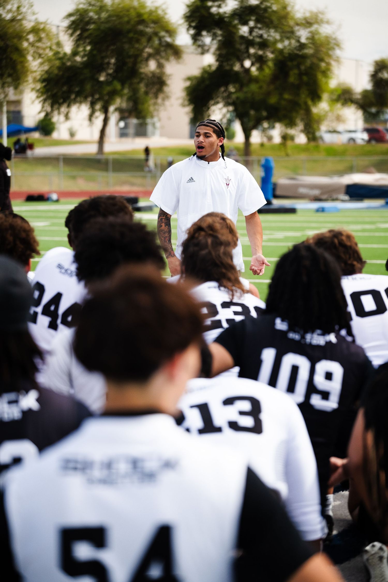 Football coach in white shirt addresses team of players in black and white uniforms on field.