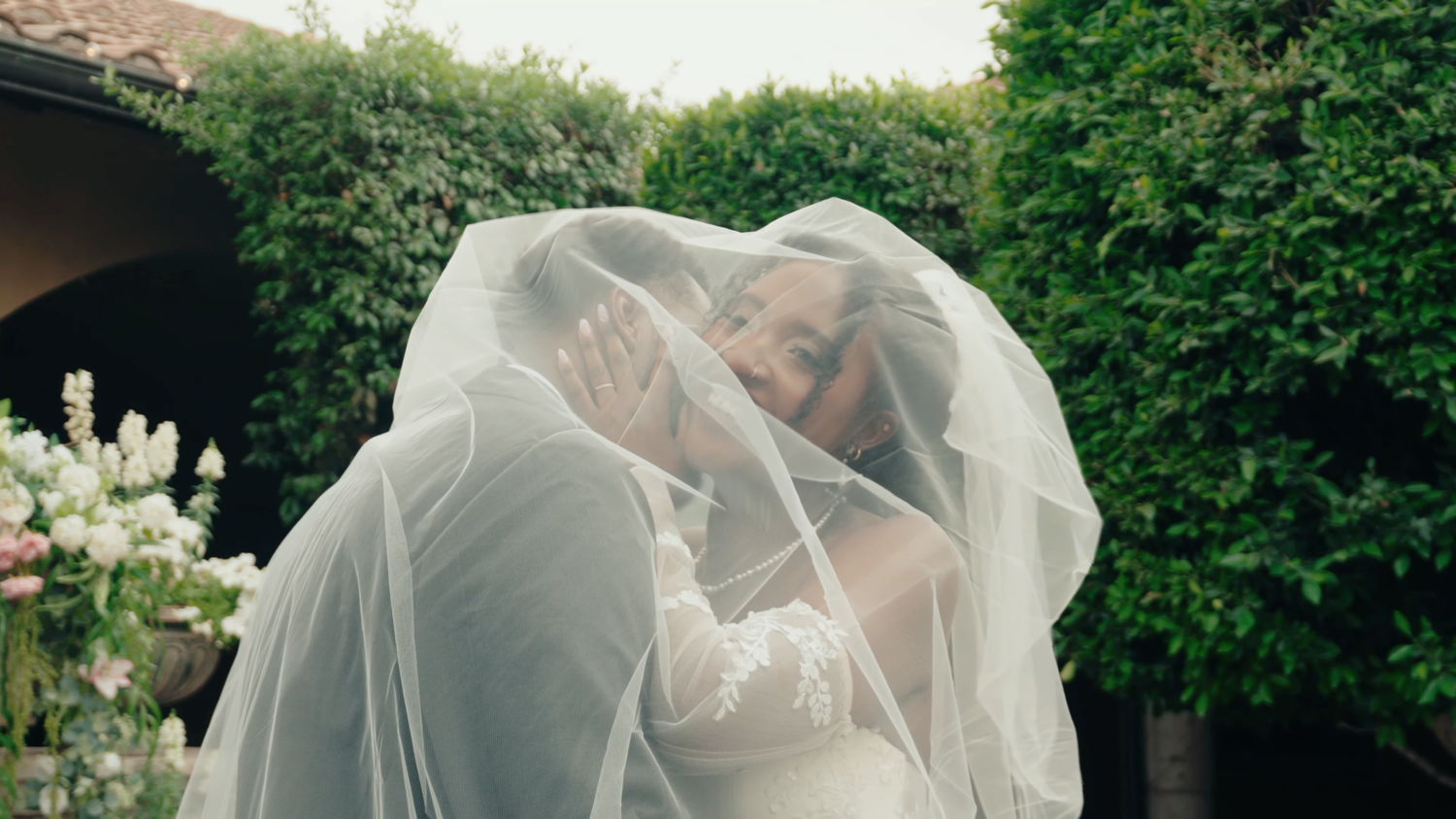 Bride and groom share romantic kiss hidden under flowing white wedding veil outdoors.