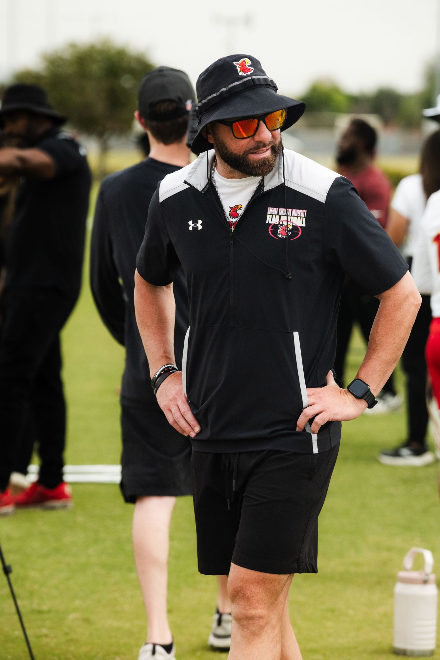 A male coach in black Under Armour athletic wear and bucket hat with sunglasses stands on a green field during football practice.