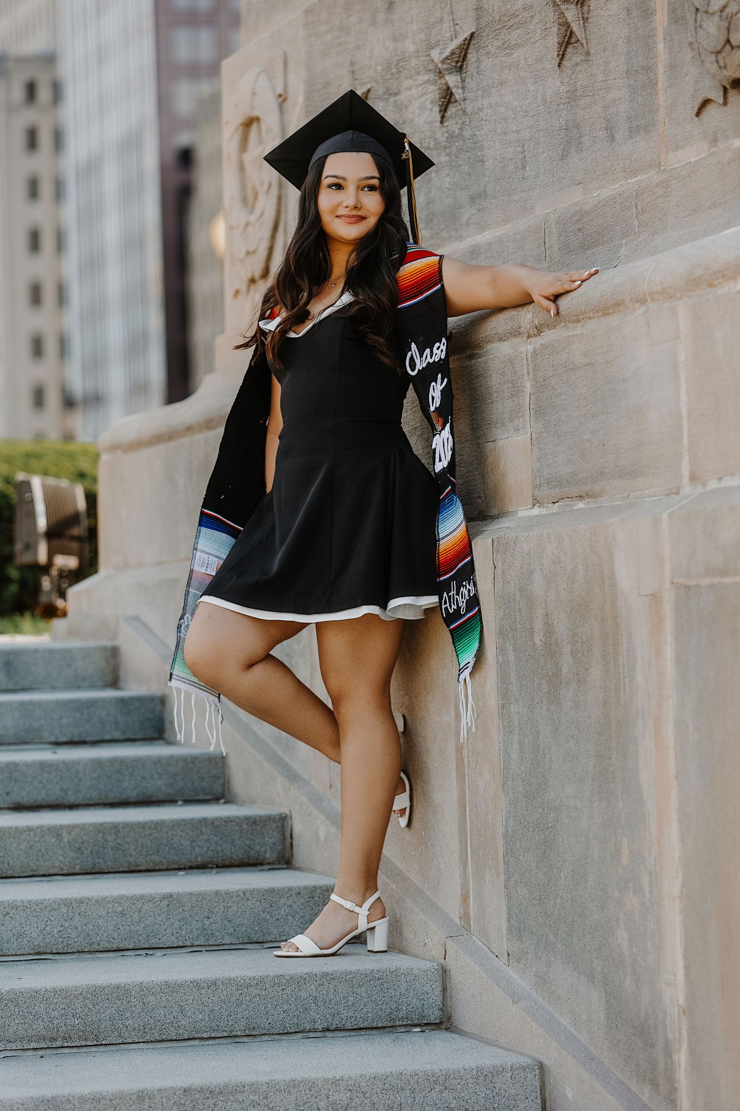 Graduate in black dress and cap with colorful sash posing on stone steps against building.