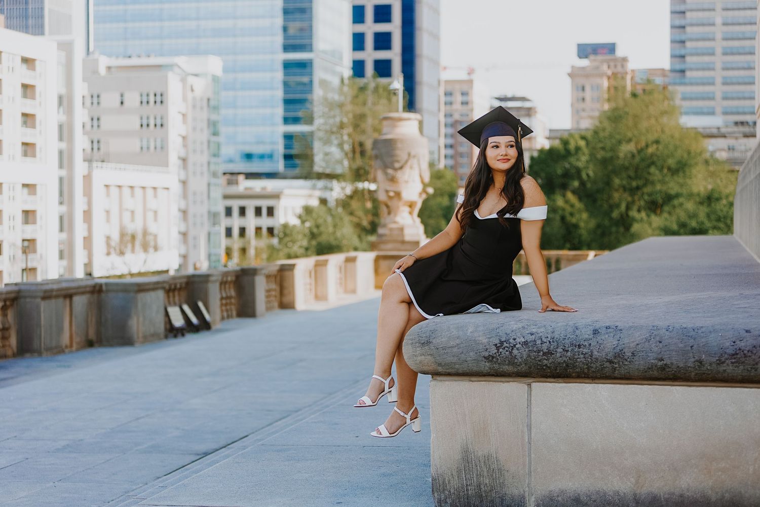 Young woman in black graduation dress and cap sitting on concrete ledge with city skyline background.