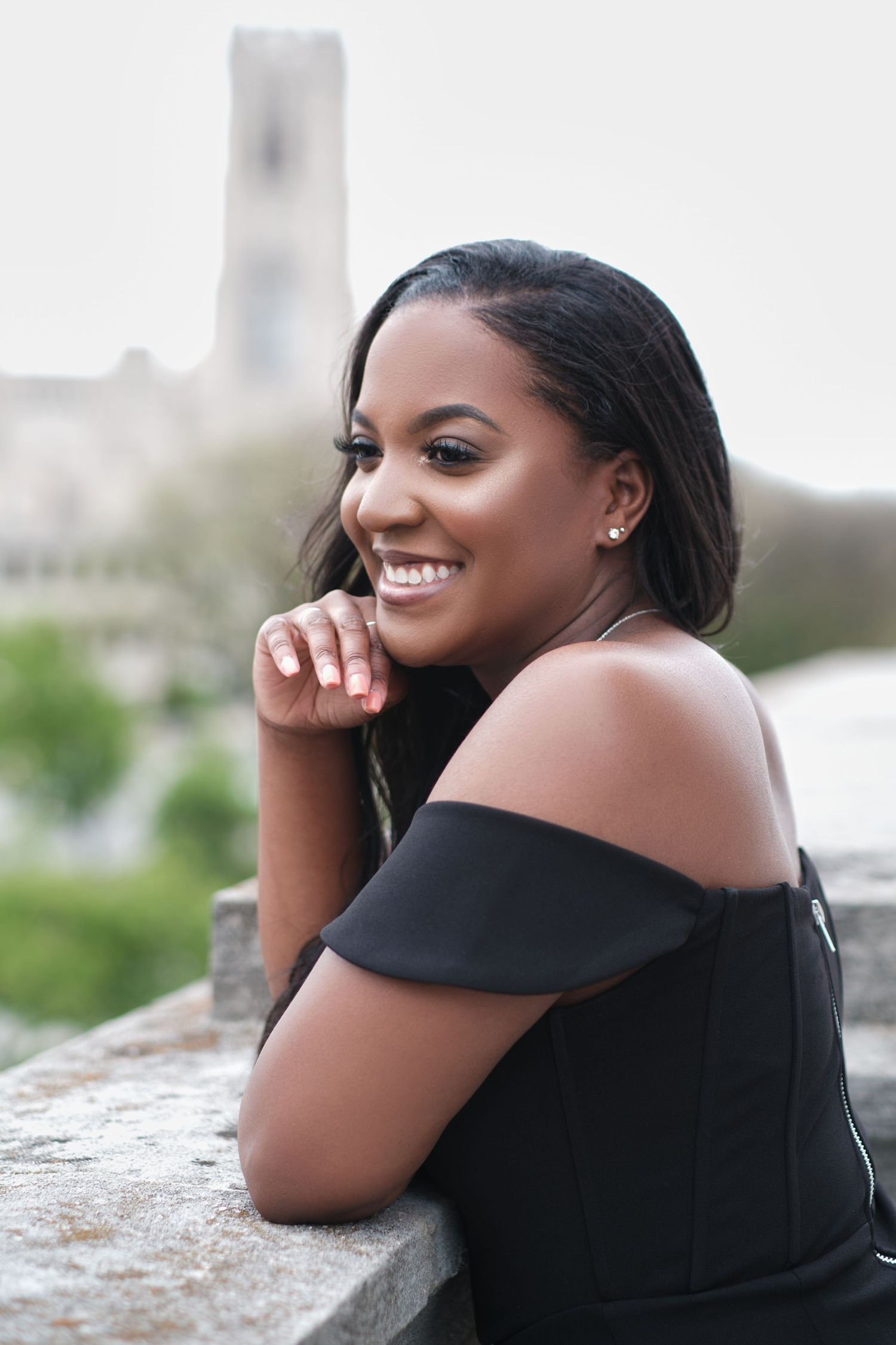 Young woman in black off-shoulder dress smiling while leaning on stone wall outdoors.