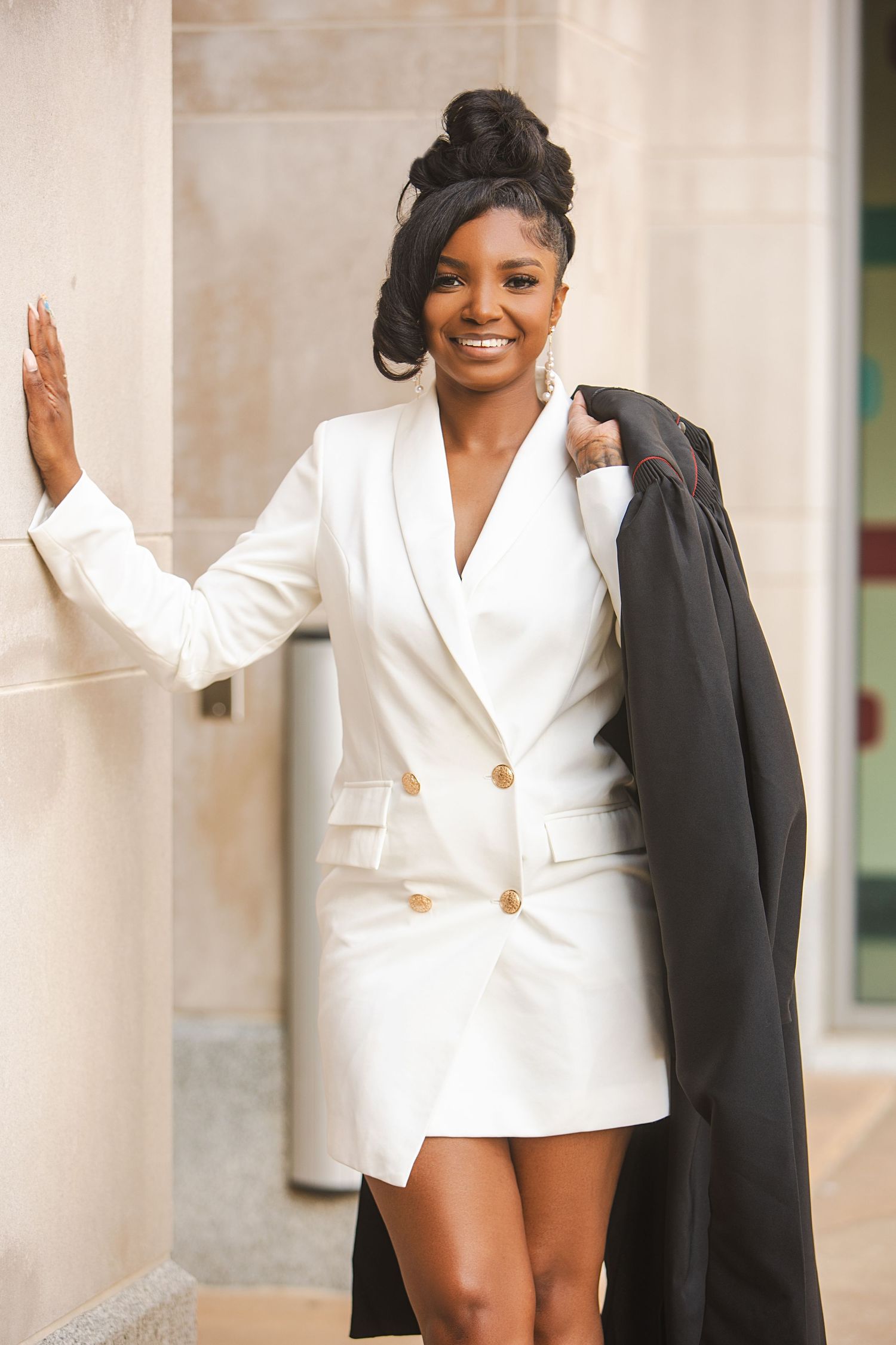 Stylish graduate in white blazer dress holds black graduation gown while leaning against stone column.