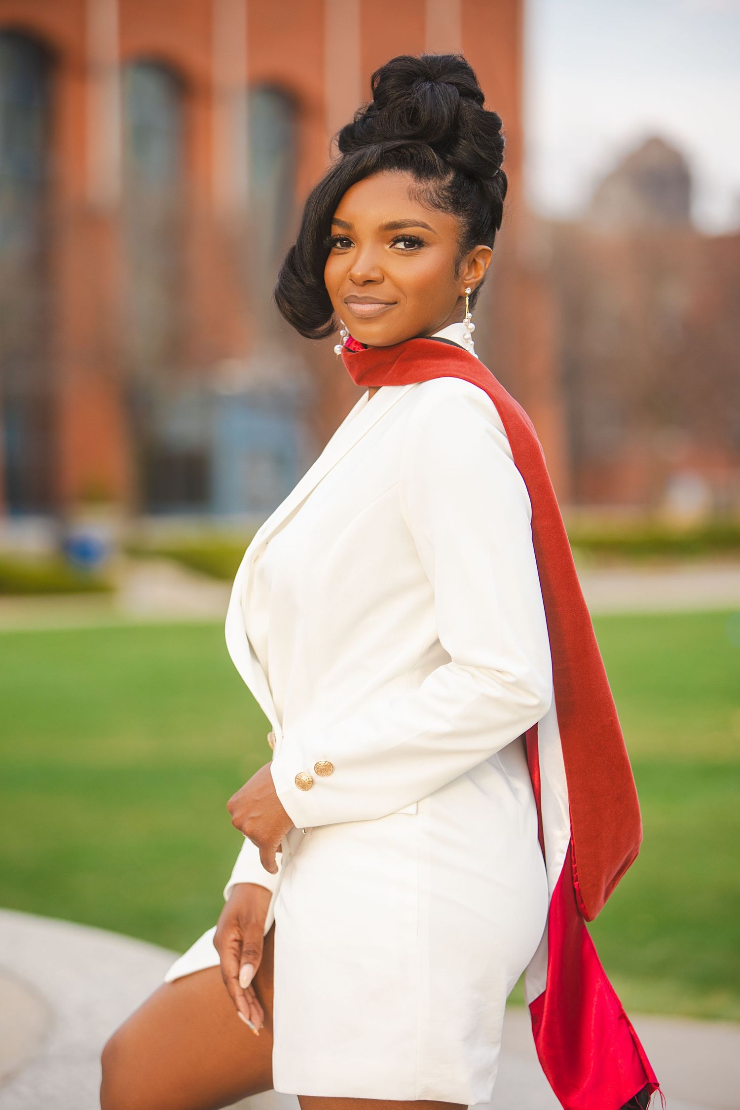 Elegant graduate in white blazer dress with red stole poses outdoors on university campus grounds.