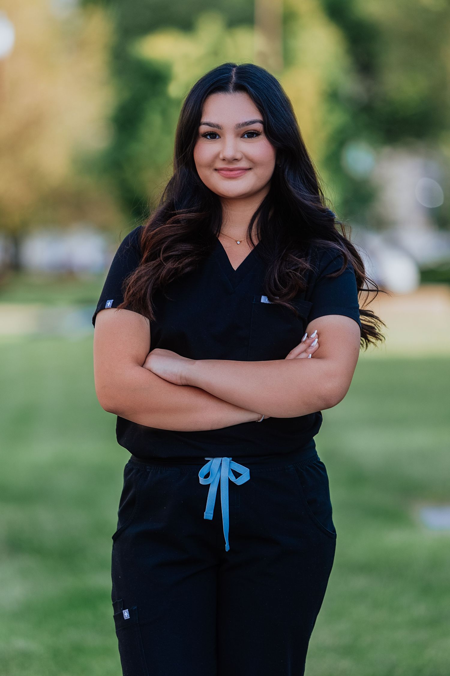 Young woman in black medical scrubs with arms crossed smiling outdoors in park setting.