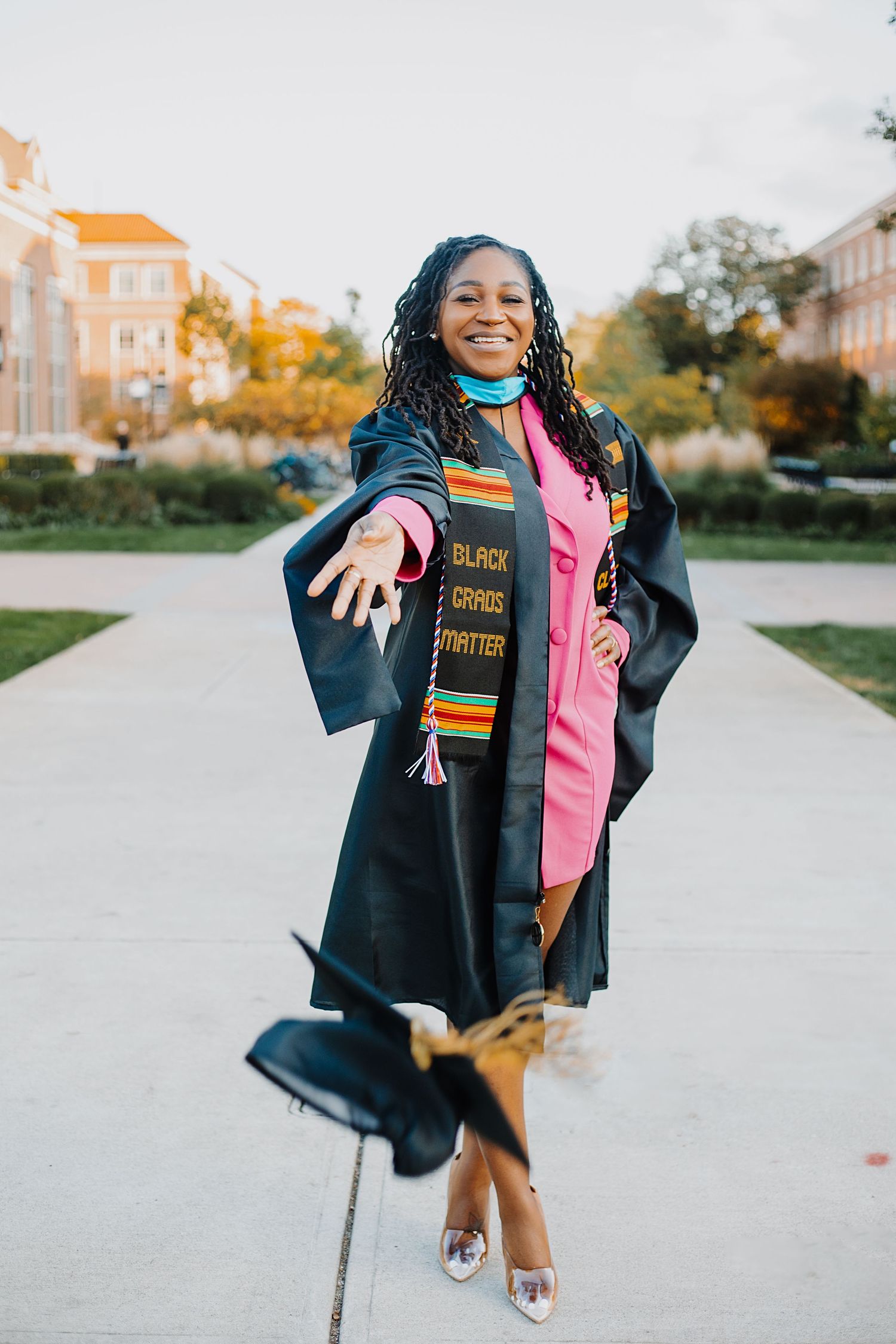 Joyful graduate in black gown with colorful stoles tosses cap on tree-lined university walkway.