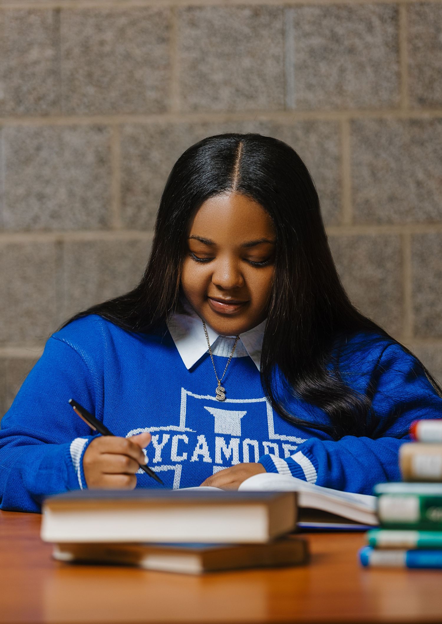 Young woman in blue Sycamore college sweatshirt studies at desk with books and school supplies.