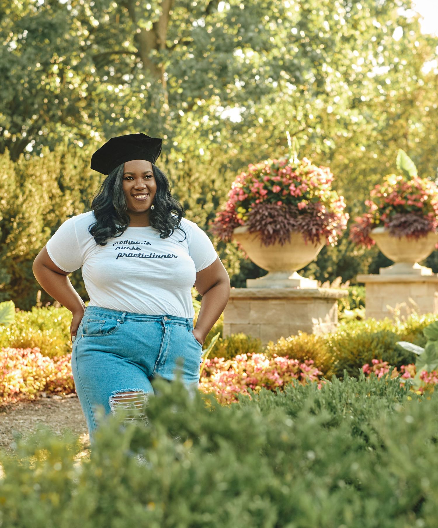 Smiling woman in white graphic tee and beret poses confidently in sunlit garden with flowers.