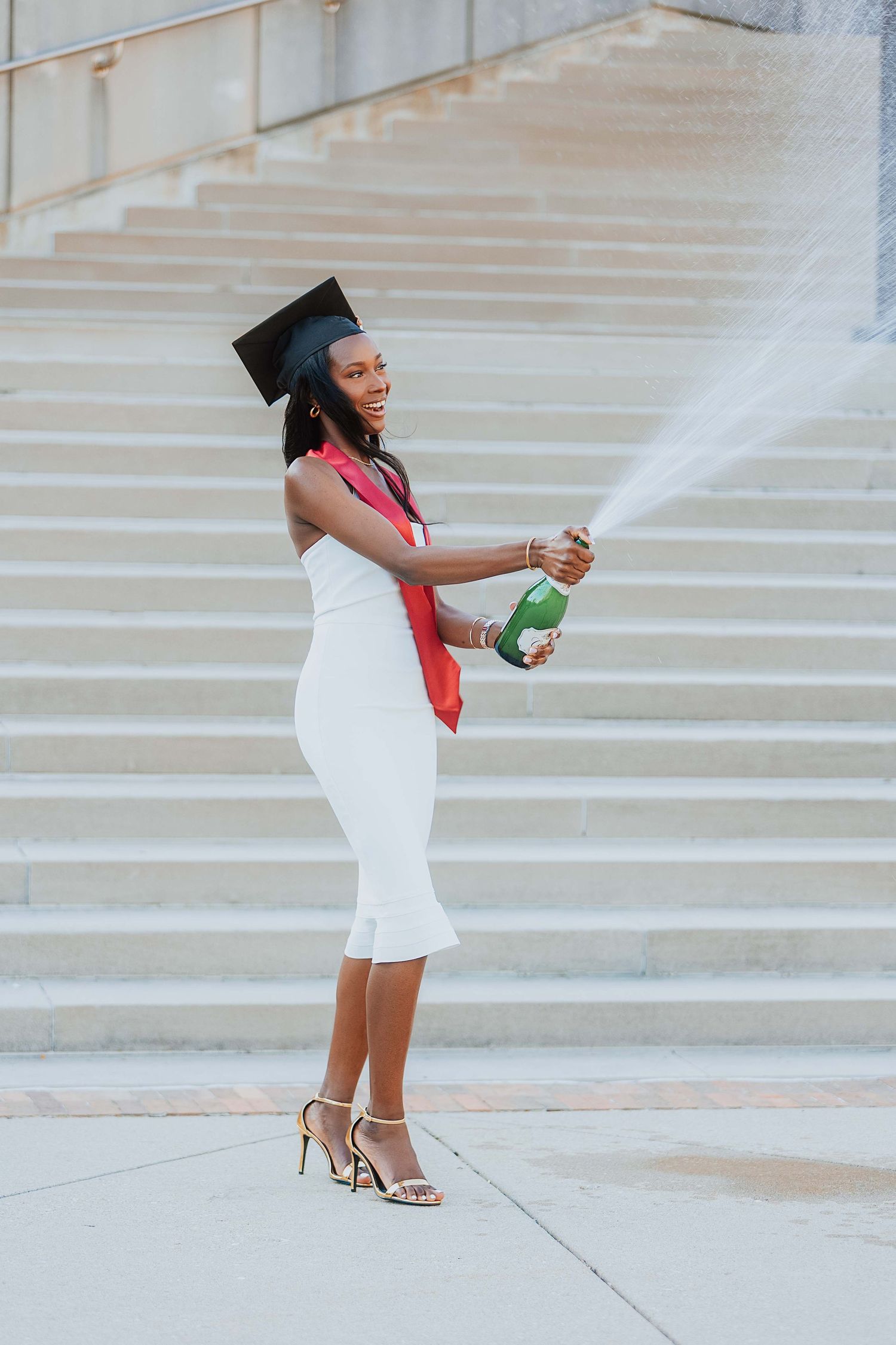 Smiling graduate in white dress and cap celebrates by spraying champagne bottle on outdoor steps.