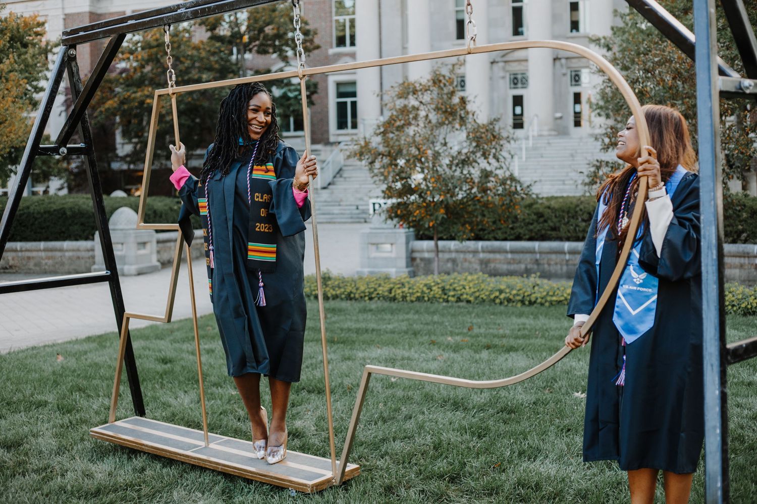 Two graduates in black gowns with colorful stoles pose near decorative swing on campus lawn.