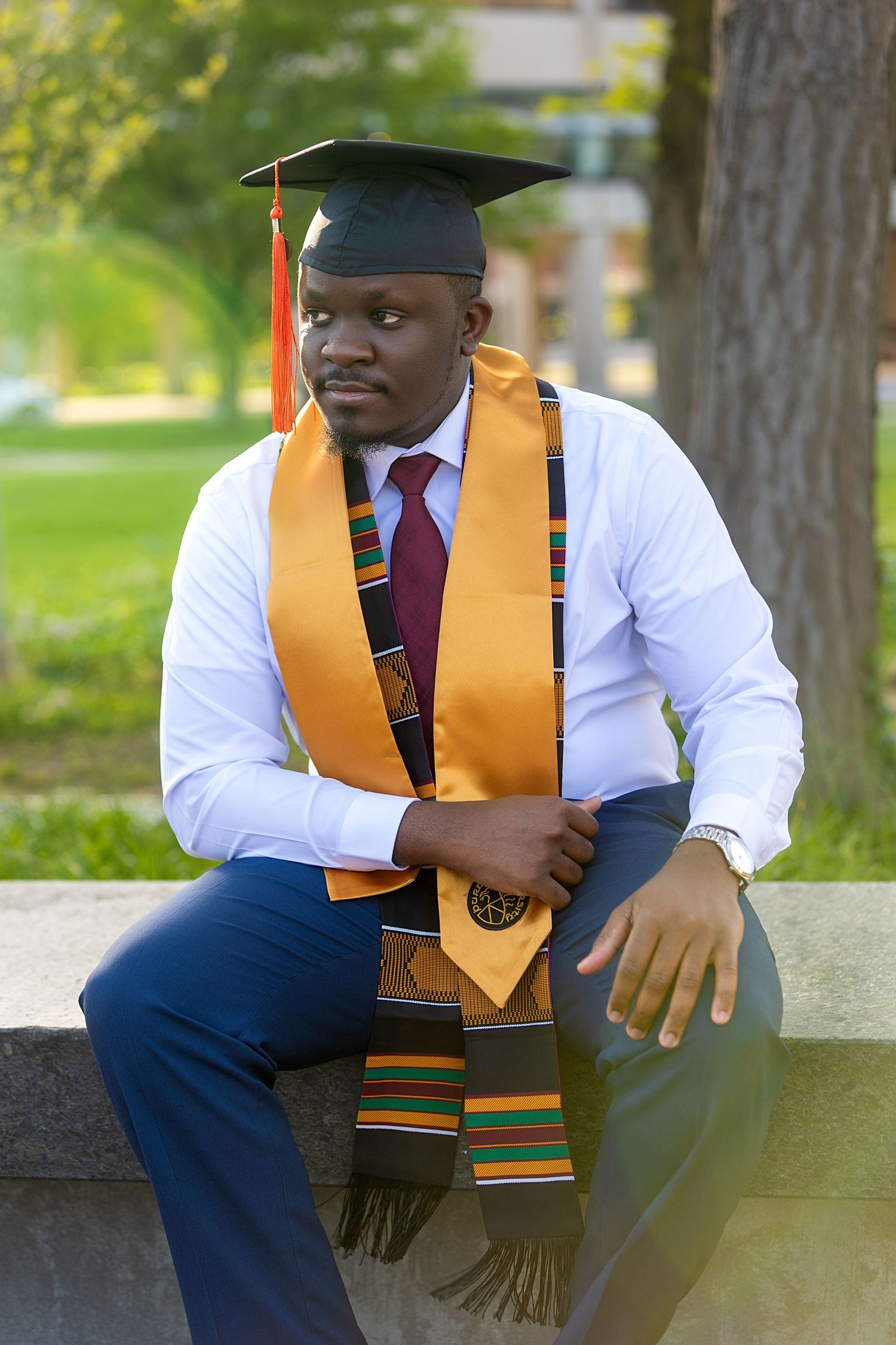 Young man in graduation cap with kente stole sitting on stone bench in park.