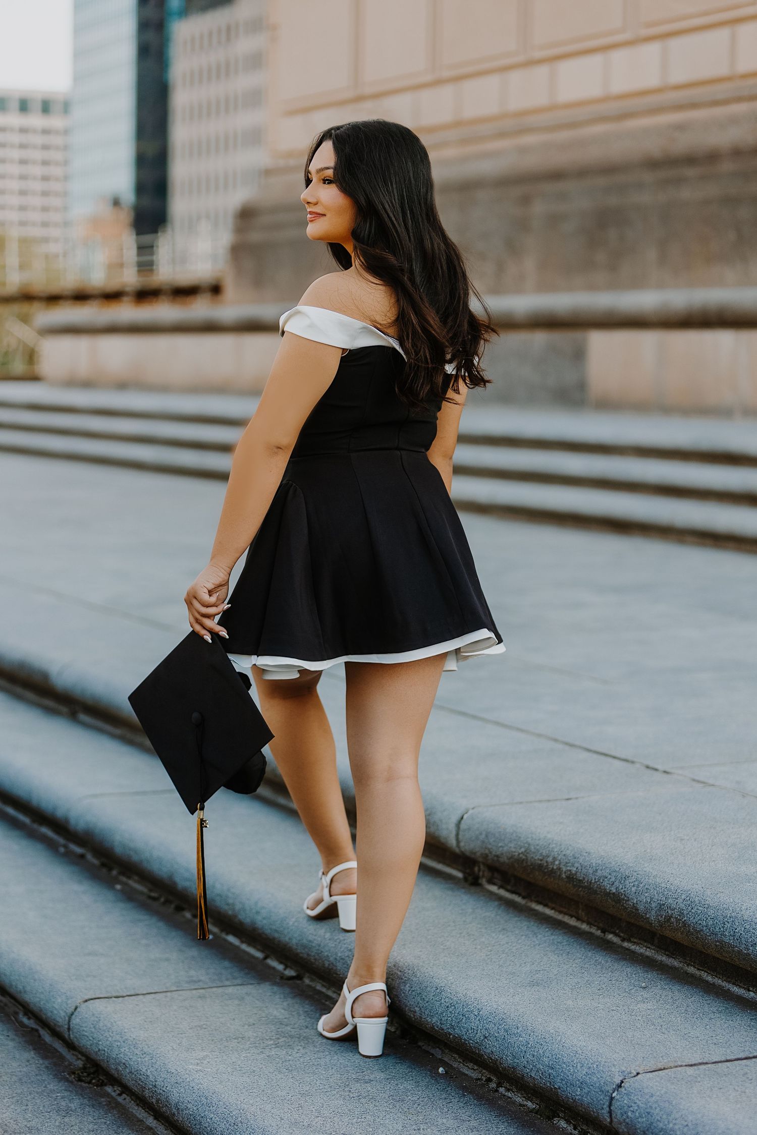 Graduate in black off-shoulder dress holds cap while posing on urban steps during graduation photo session.