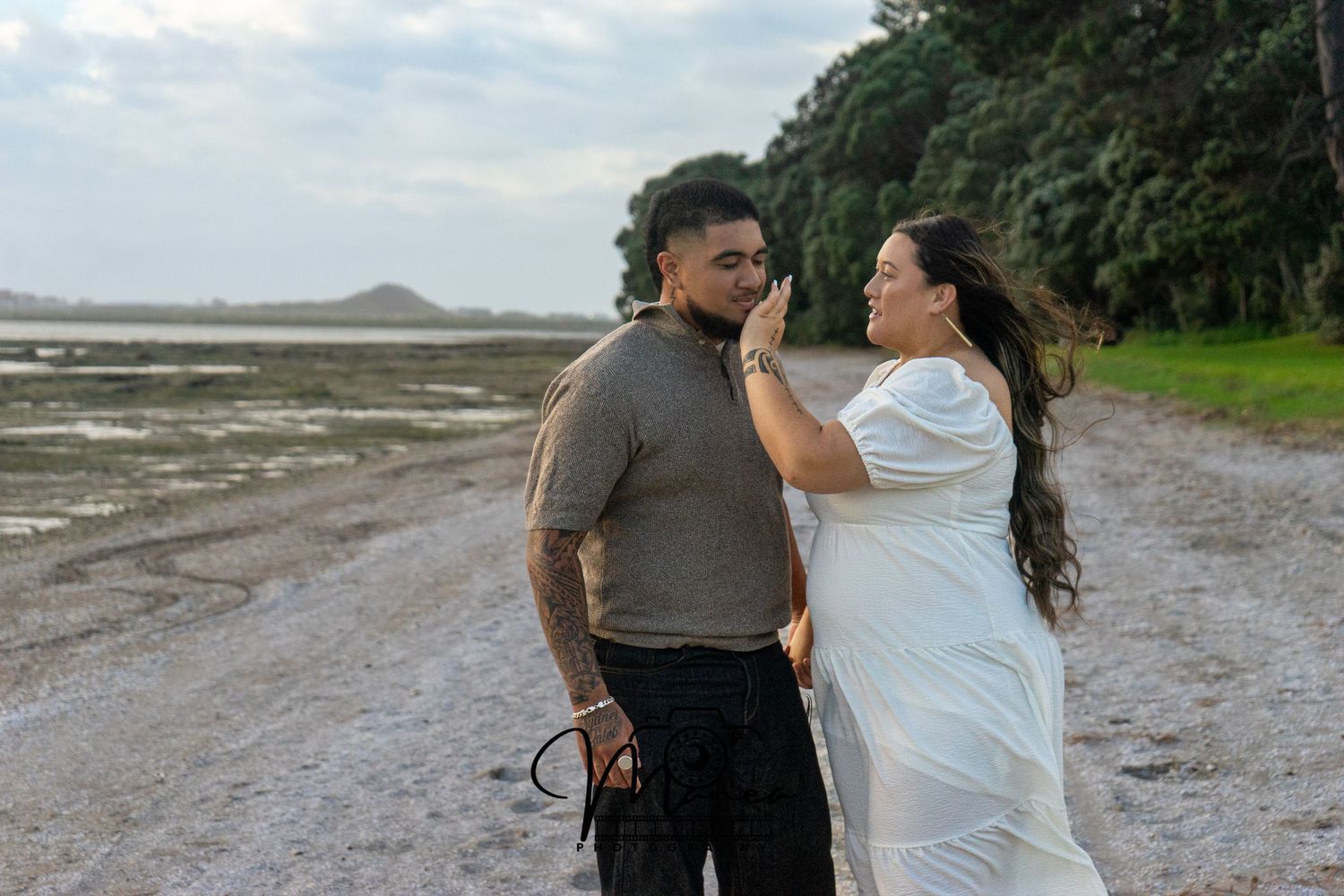 Couple sharing a tender moment on a coastal beach path with lush green trees in the background.